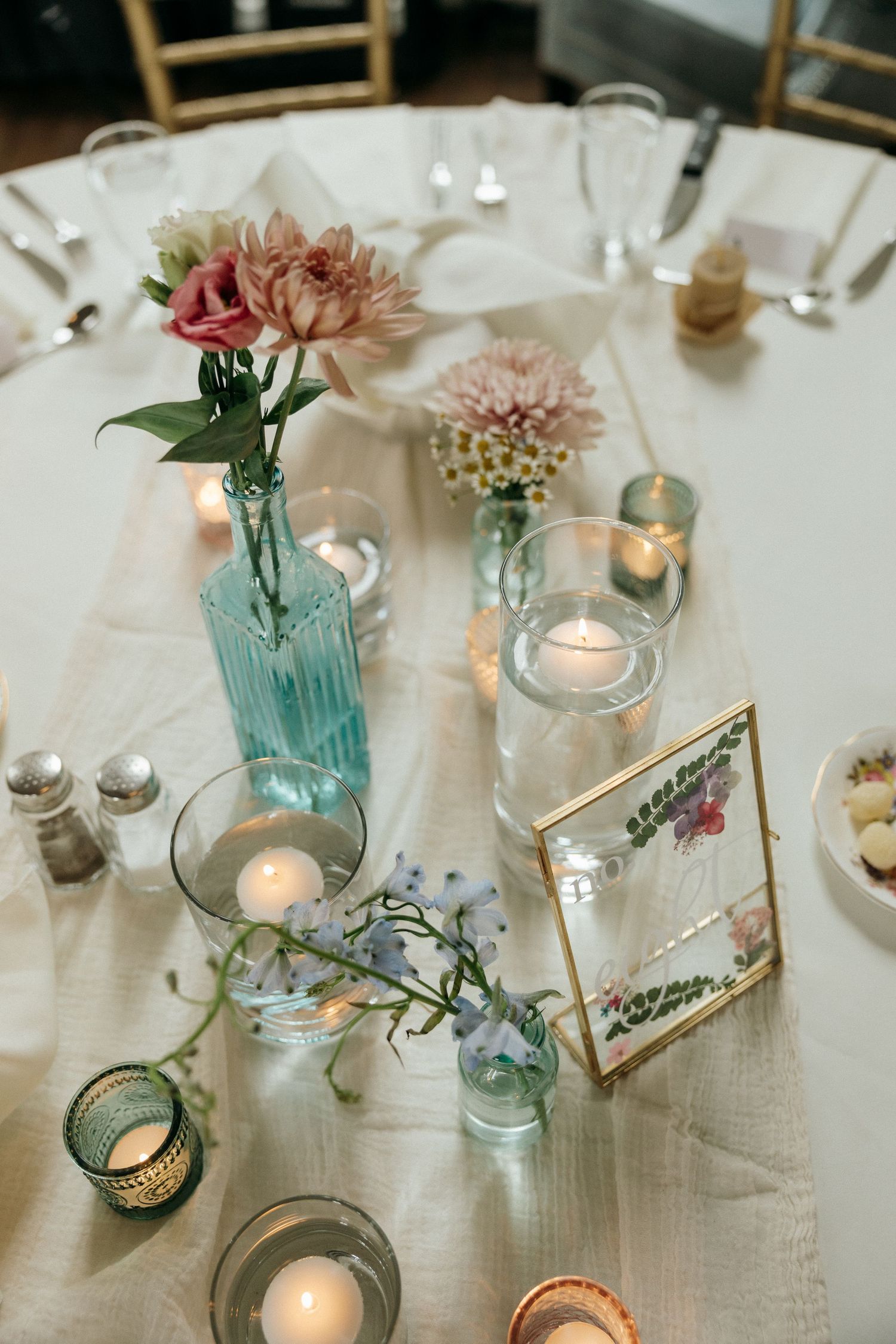 Elegant wedding table decor with blue glass vases, delicate flowers, and glowing candles on white linens.