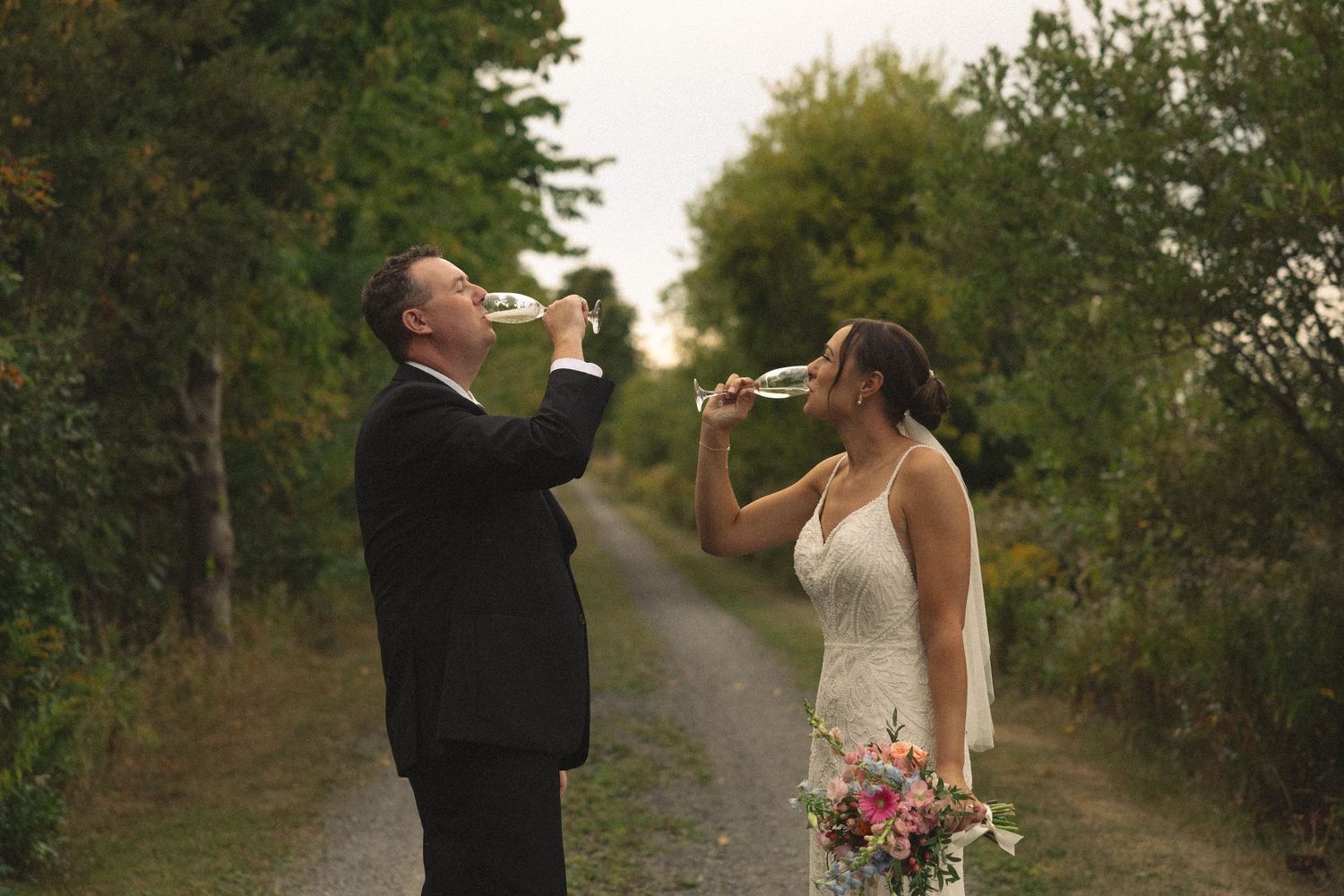 Playful moment of couple drinking champagne on a tree-lined path with wedding bouquet.