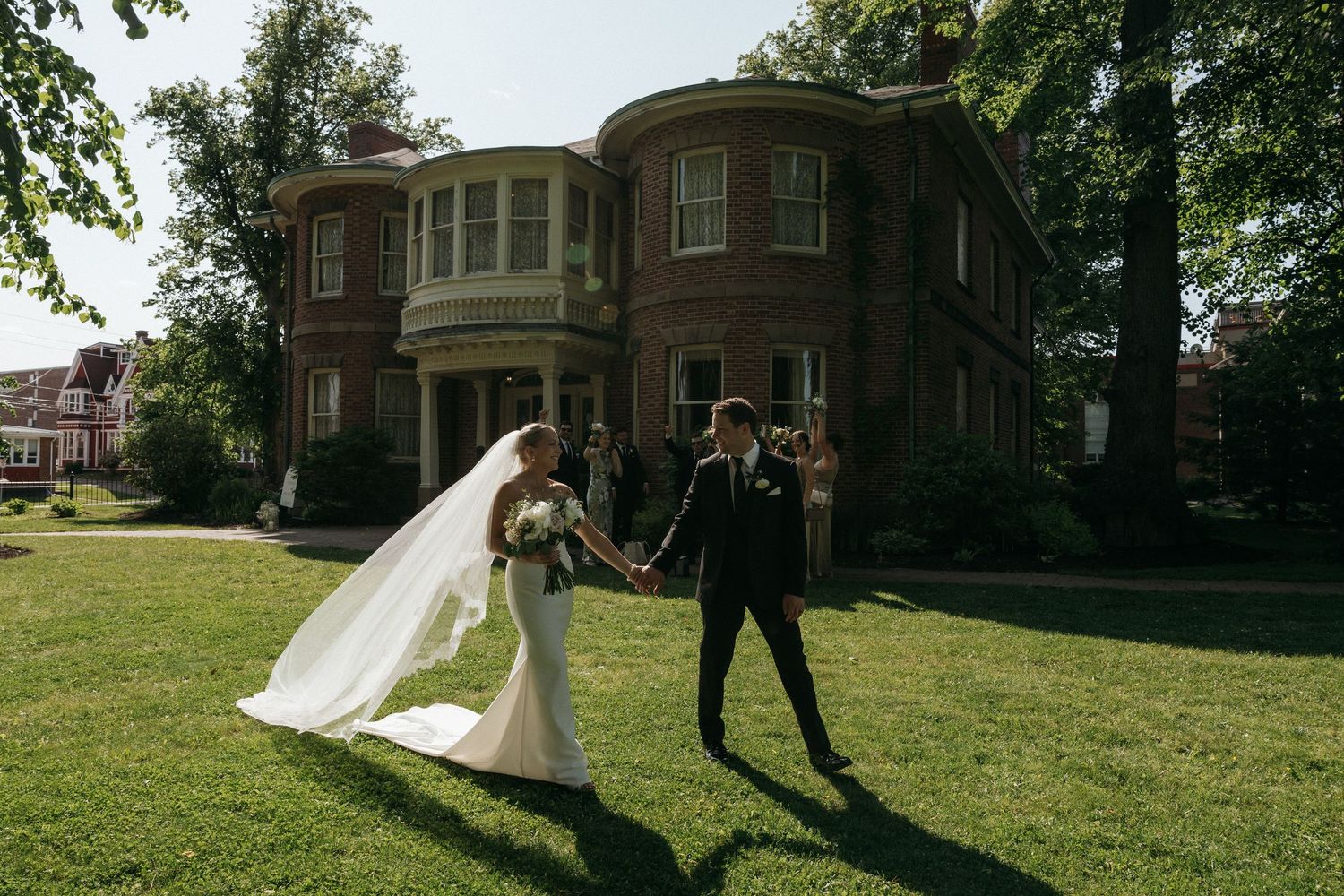 Joyful wedding couple dancing on lawn in front of historic brick mansion.