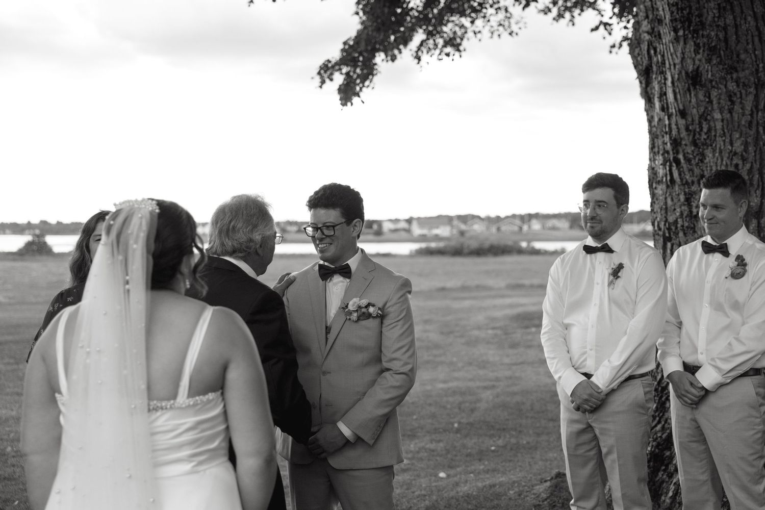 Black and white photo of bride and groom exchanging vows with officiant under a tree outdoors.