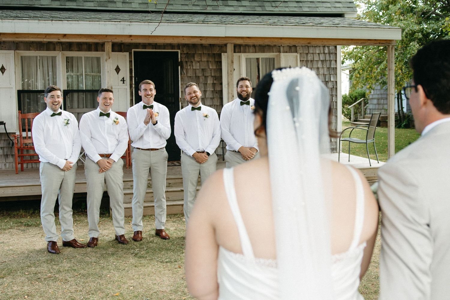 Groomsmen in white shirts and gray pants watch as bride and groom exchange vows outside rustic building.