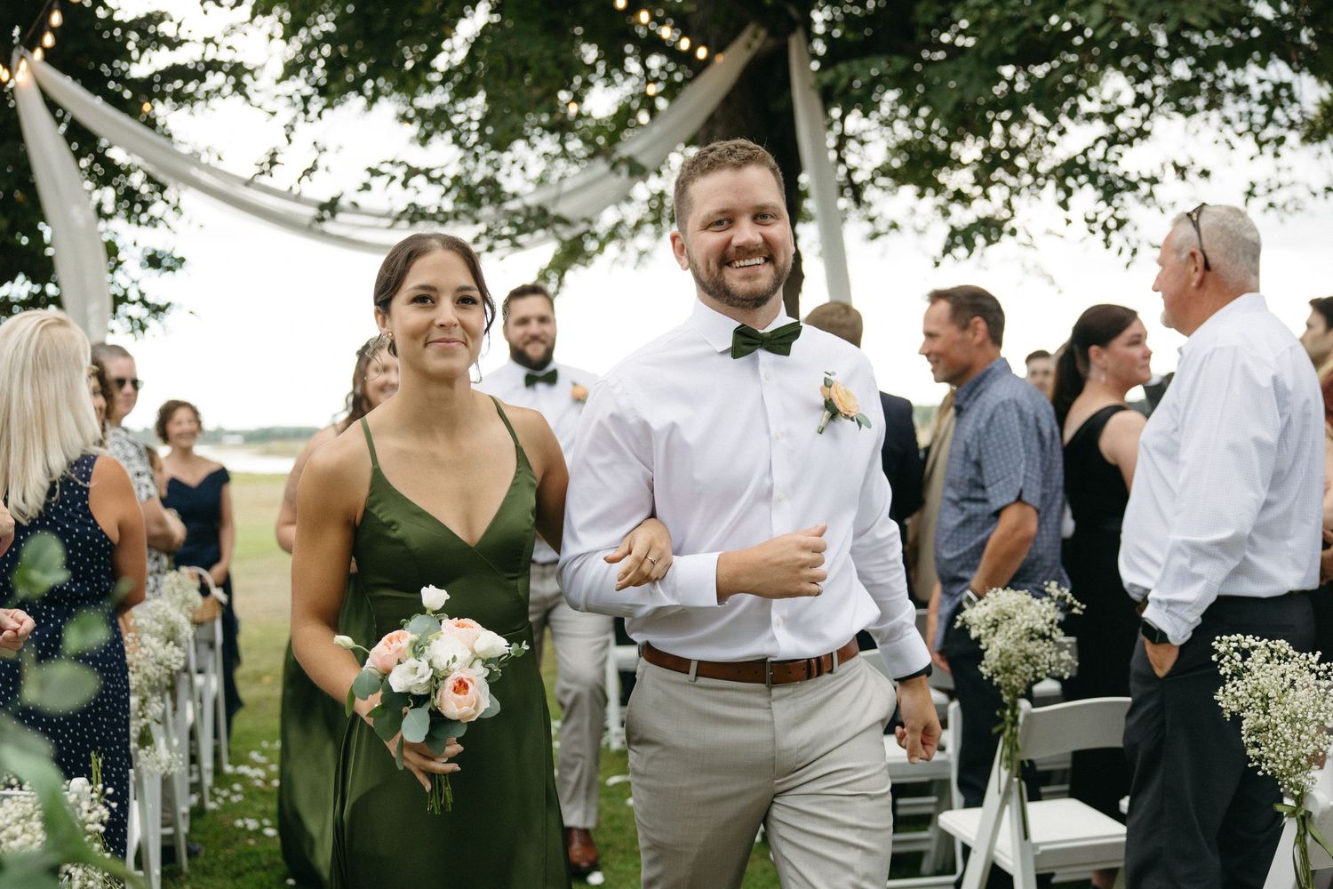 Bridesmaid in green dress walks down aisle with smiling groomsman in bow tie at outdoor wedding.
