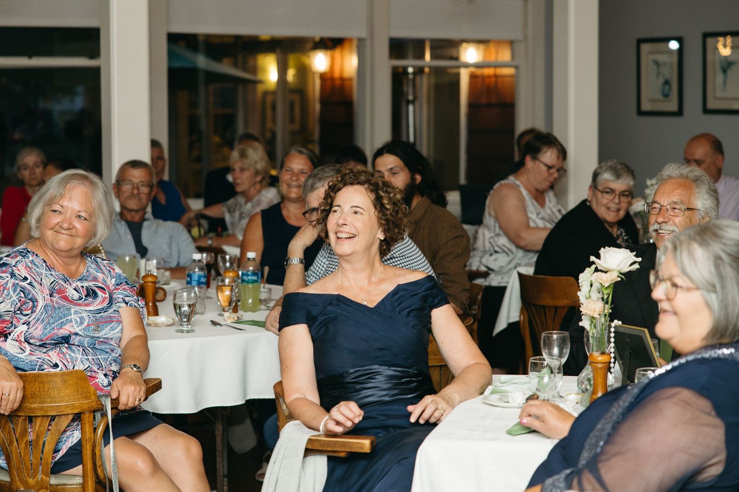 Elegantly dressed wedding guests smile and laugh during indoor reception speeches at round tables.