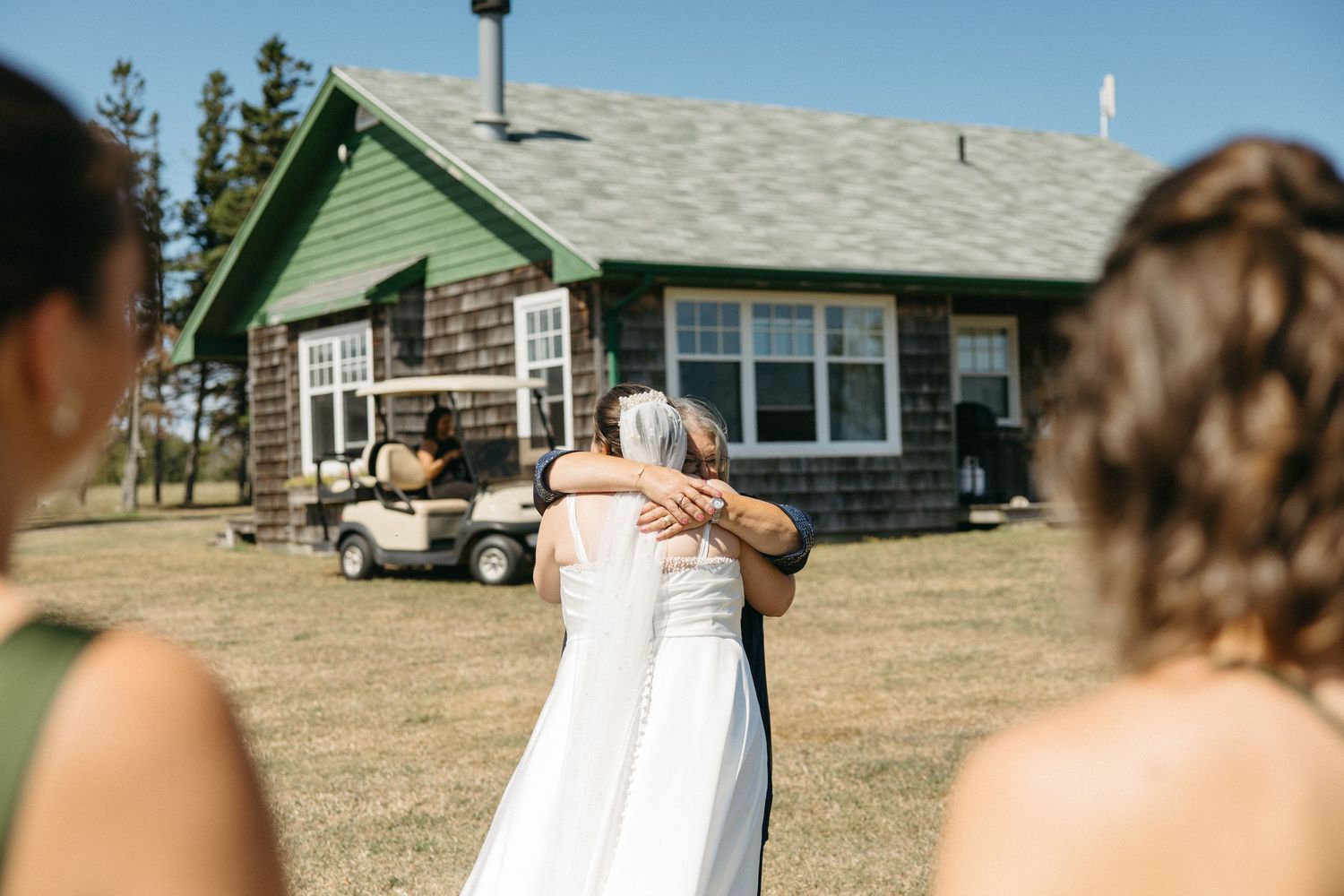 Bride in white wedding dress sharing a warm hug with a guest outside a rustic green farmhouse on a sunny day.