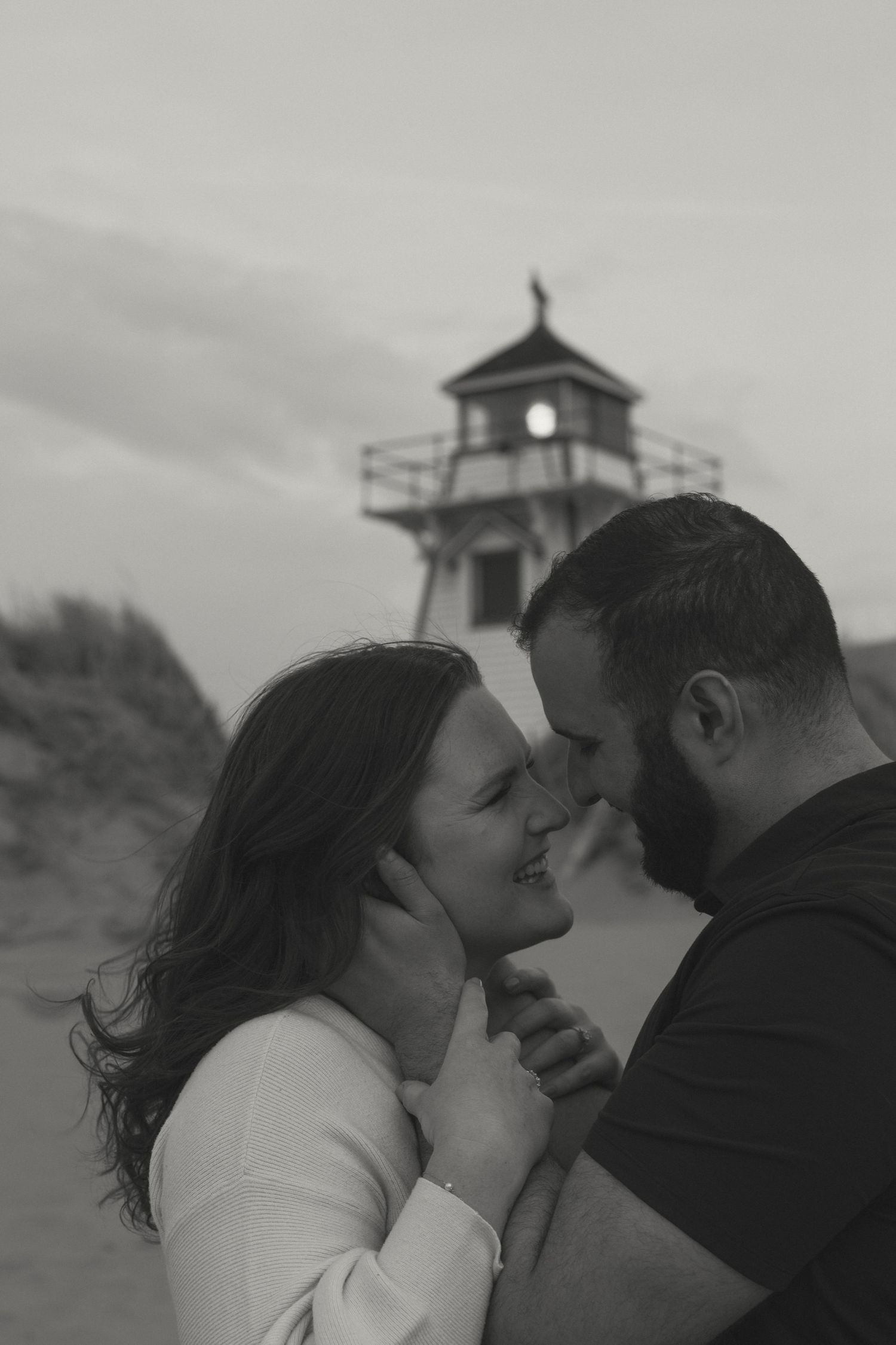 A couple shares an intimate moment on a beach with a lighthouse visible in the background.