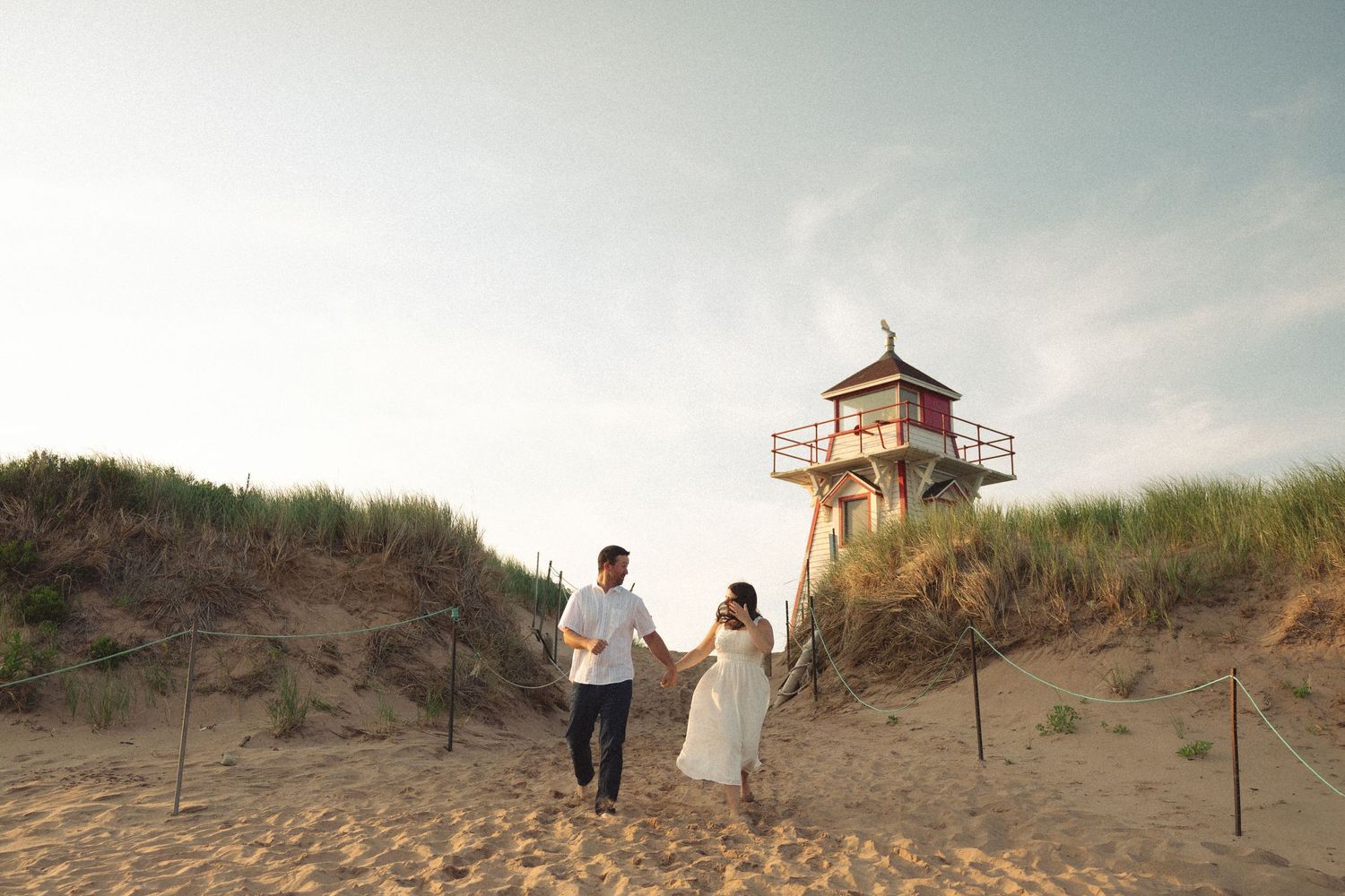 Wide angle view of a lighthouse and couple walking on sandy beach during golden hour.