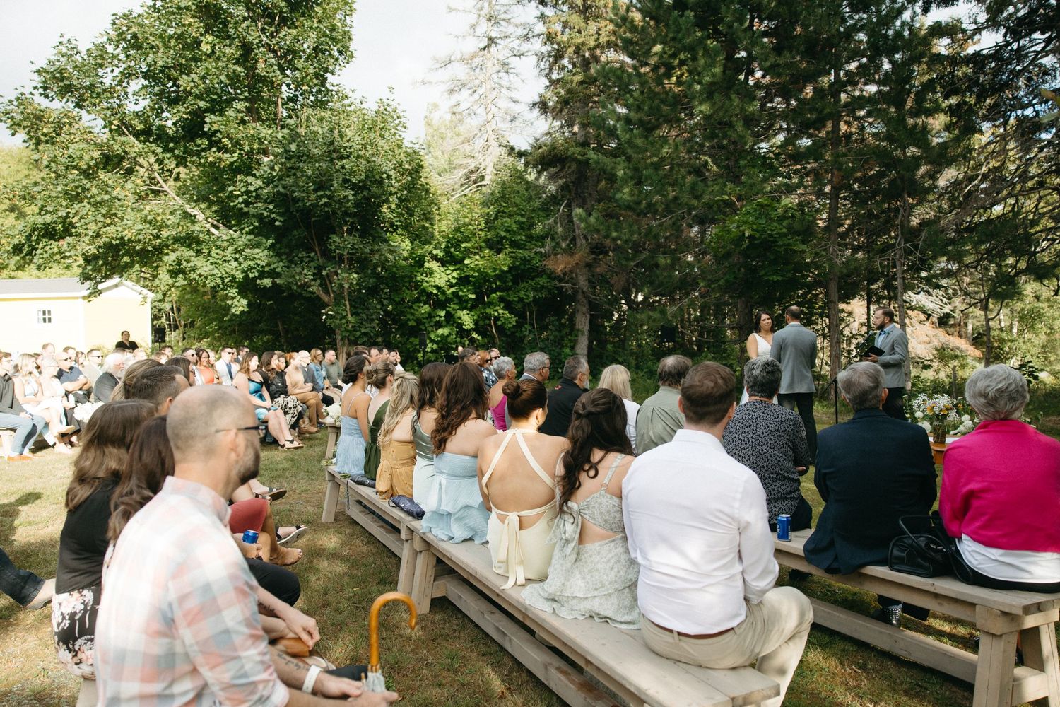 Guests seated on wooden benches at an outdoor ceremony by a lake surrounded by evergreen trees.