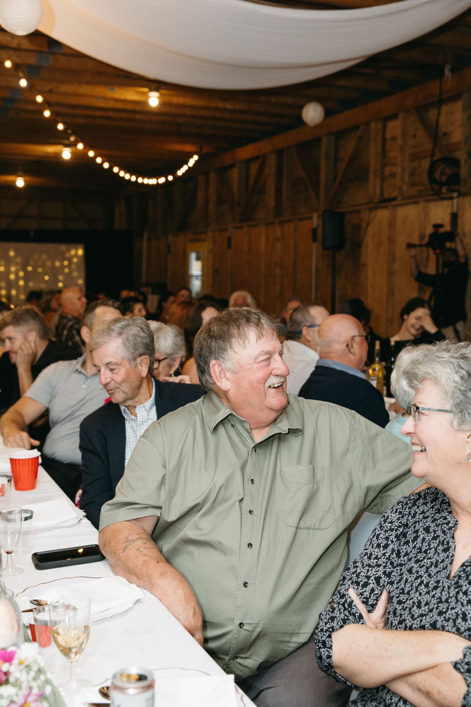 Wedding guests laugh and socialize at decorated tables during a rustic barn reception.