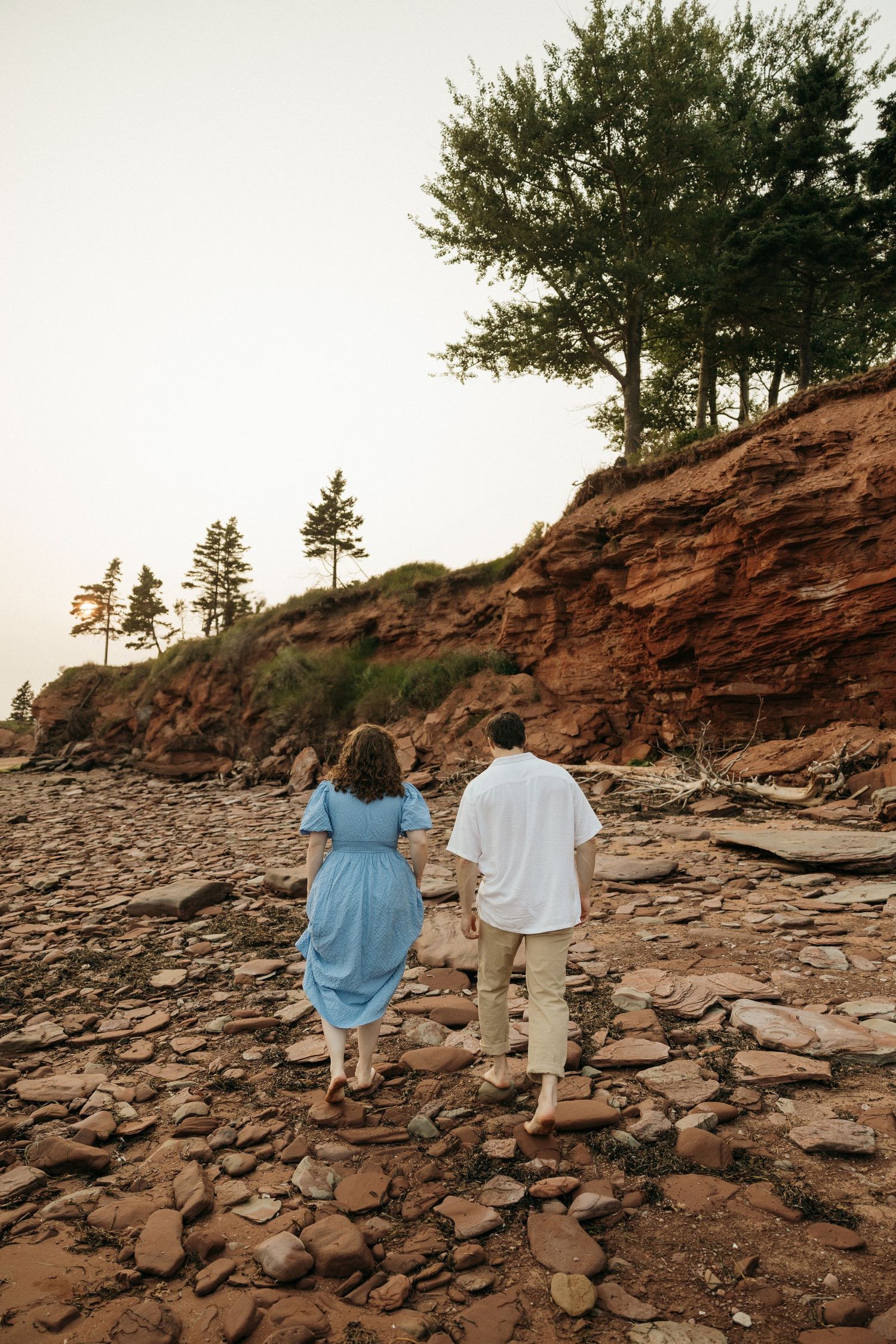 Couple walking together on rocky terrain near red cliffs during a sunset photoshoot.