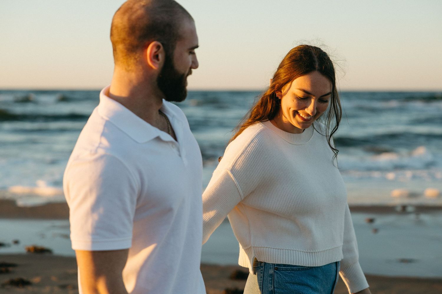 Couple in white outfits walking together along the ocean shore at sunset.
