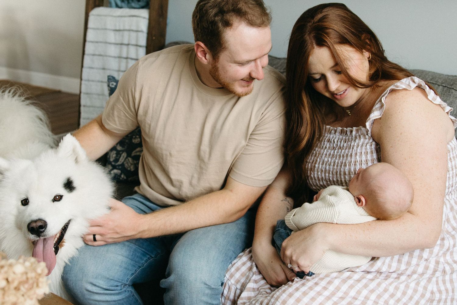 A family cuddles together on a couch with their fluffy white dog in natural light.