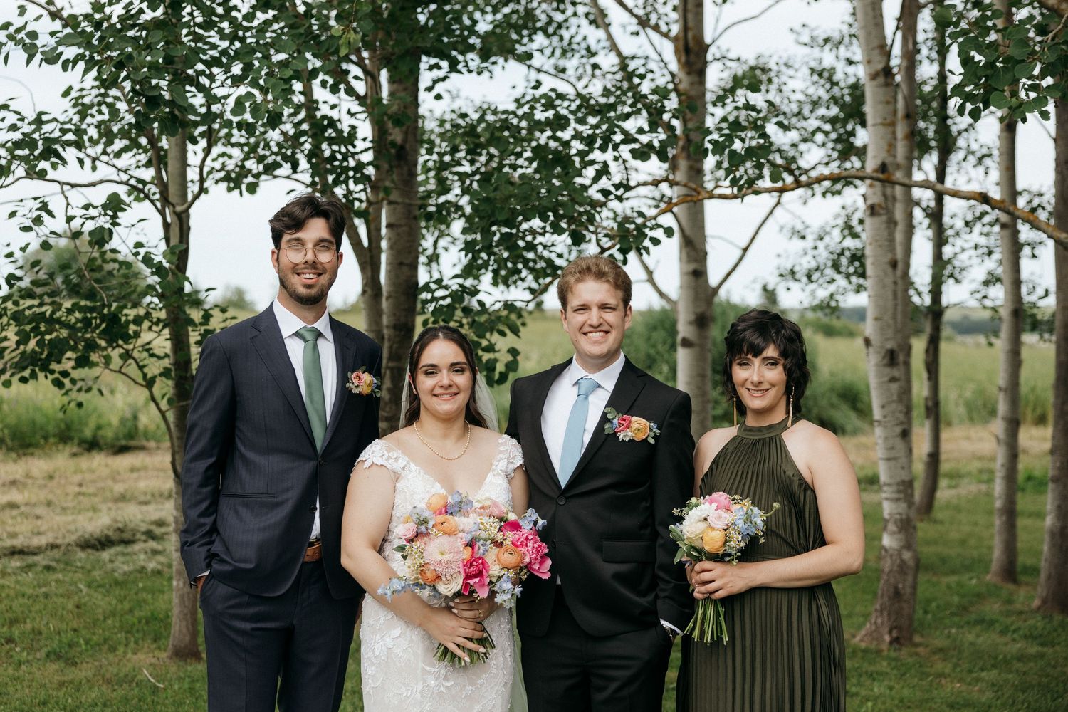 Wedding group standing among trees holding vibrant floral bouquets.