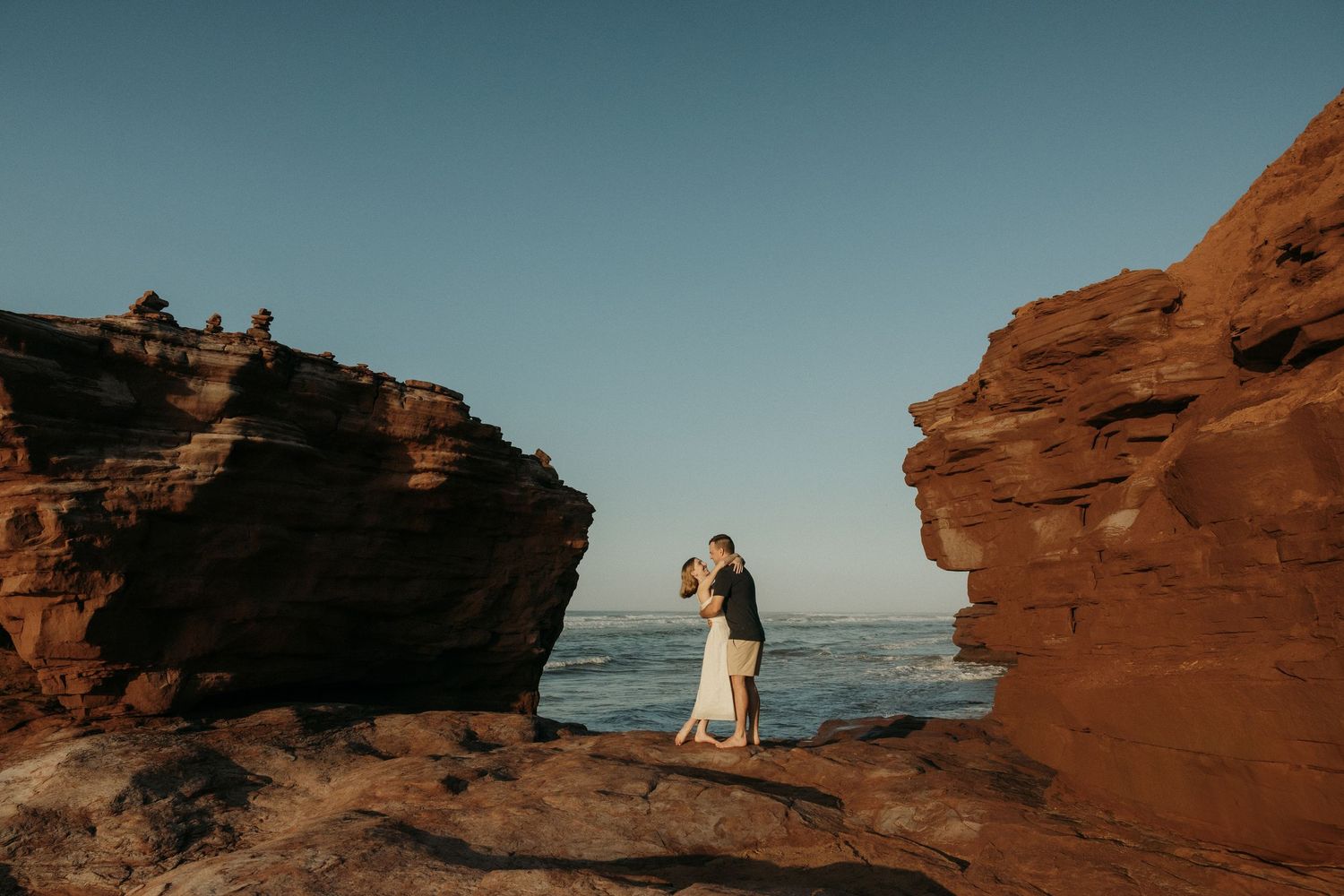 Red coastal cliffs frame a scenic ocean view at sunset with two people in the distance.