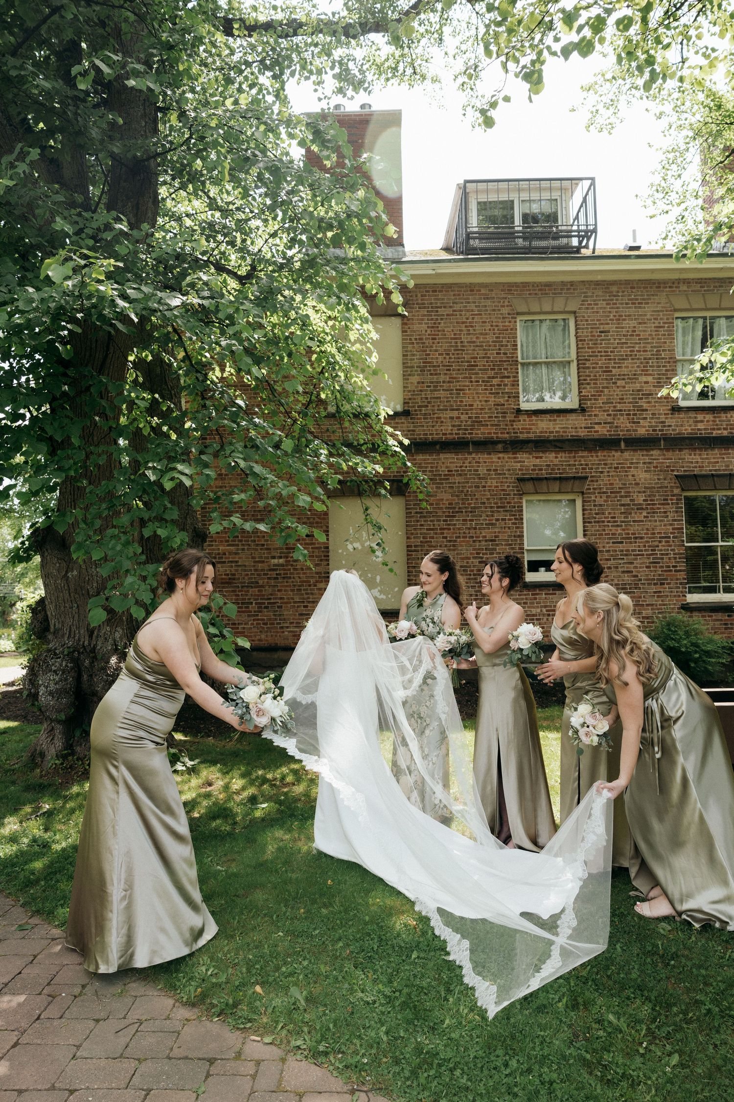 Wedding party playfully adjusts bride's flowing veil on grassy lawn near brick building.