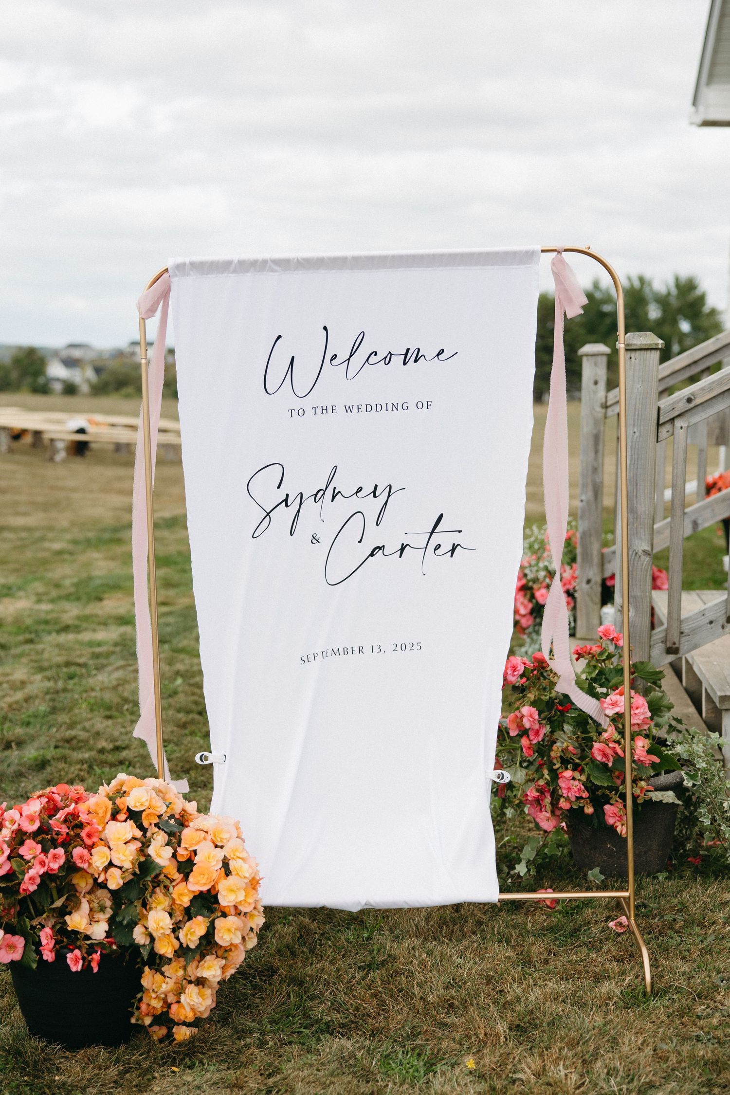 Wedding welcome sign with pink floral decorations displayed on a white banner outdoors.
