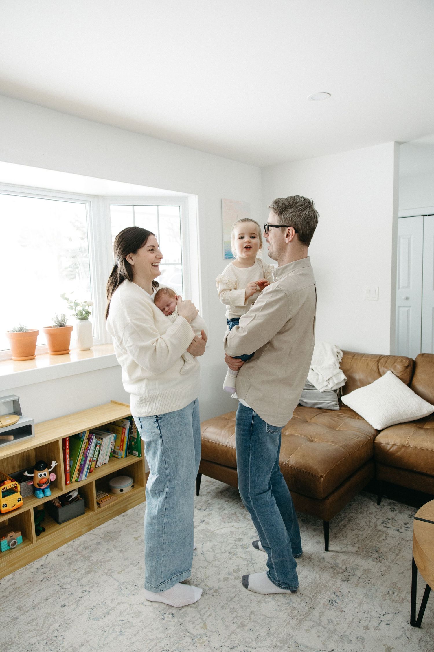 Family in matching white sweaters and jeans playing together in a minimalist living room with bookshelf.