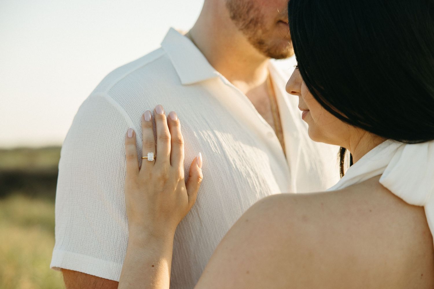 Close-up of a woman placing her hand on a man's shoulder, wearing an engagement ring, in a sunny outdoor setting.