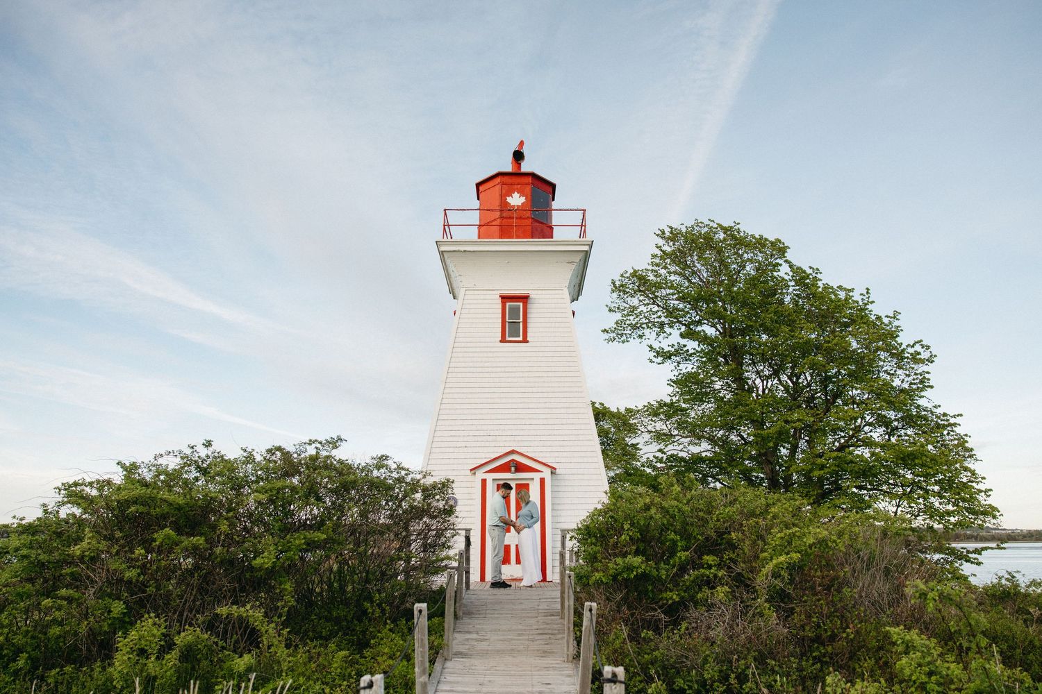 Classic white and red lighthouse surrounded by lush green trees under a partly cloudy blue sky.