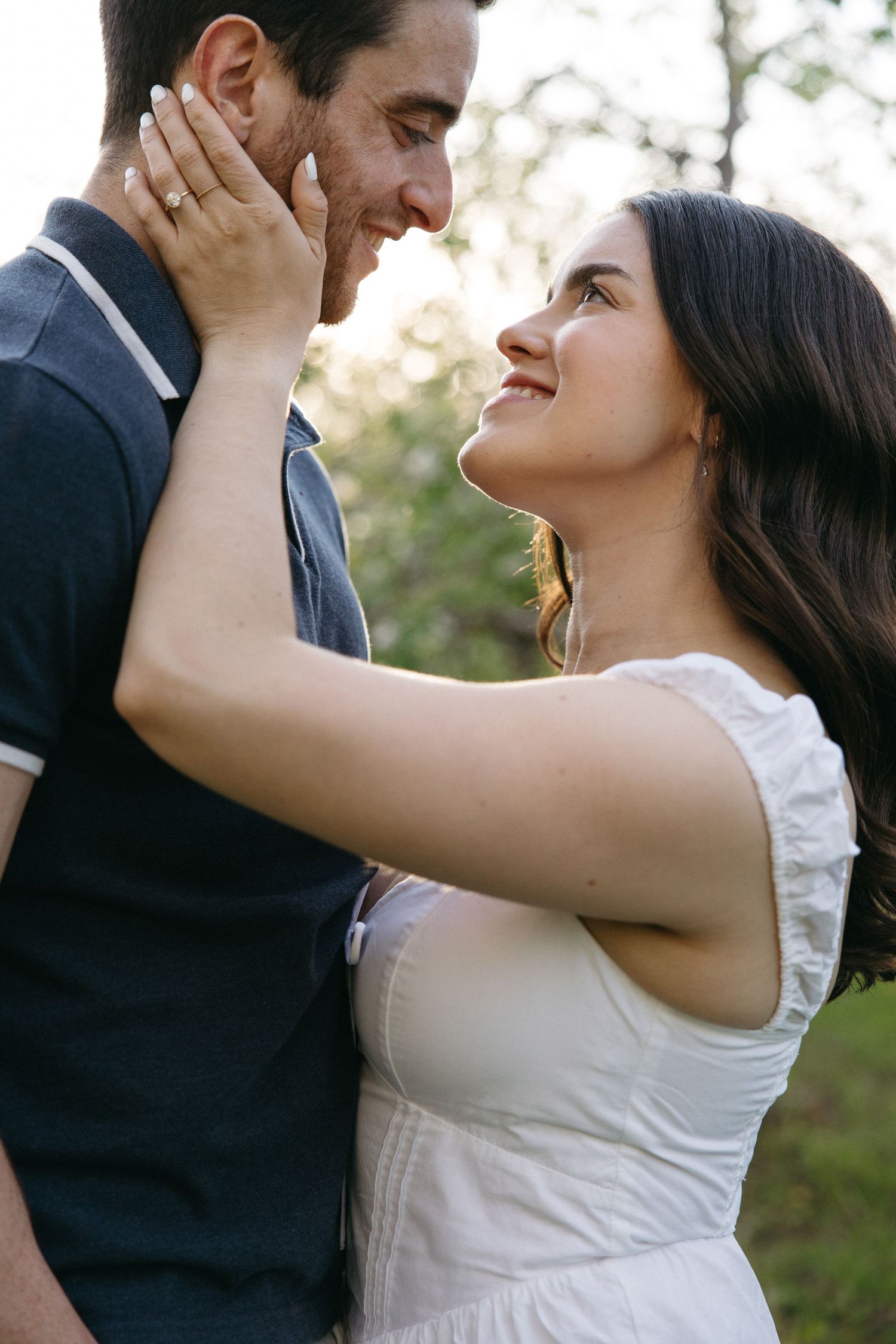 A smiling couple shares a tender moment outdoors at sunset, woman in white dress gently holding mans face.