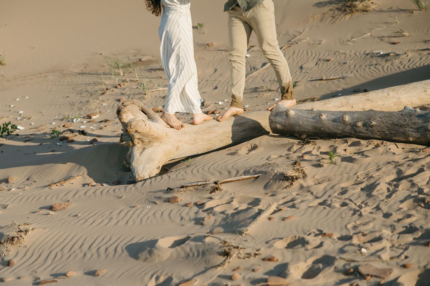 Couple walking barefoot on sandy beach near driftwood log in neutral toned clothing.