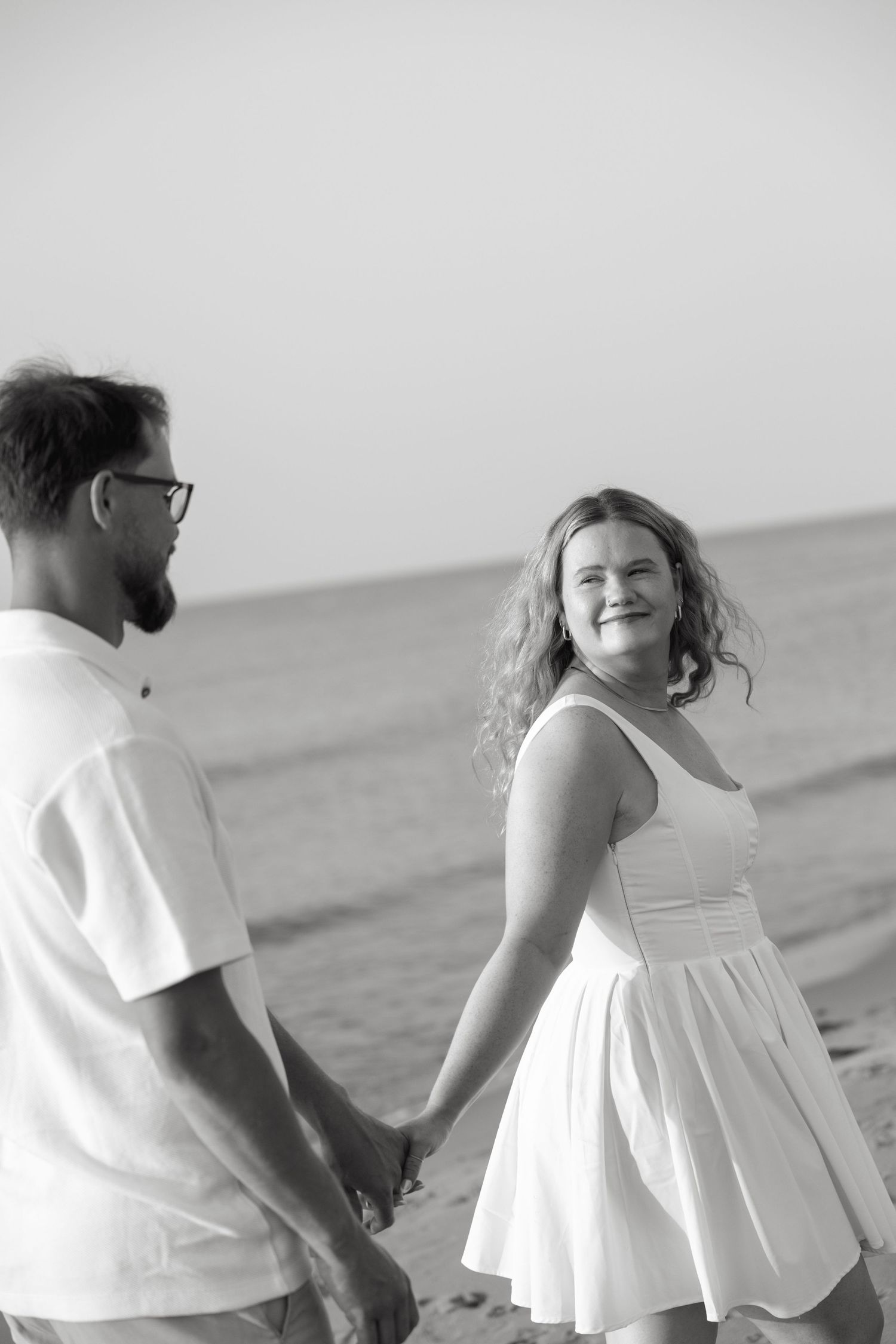 Black and white photo of couple holding hands on beach, woman smiling over her shoulder.