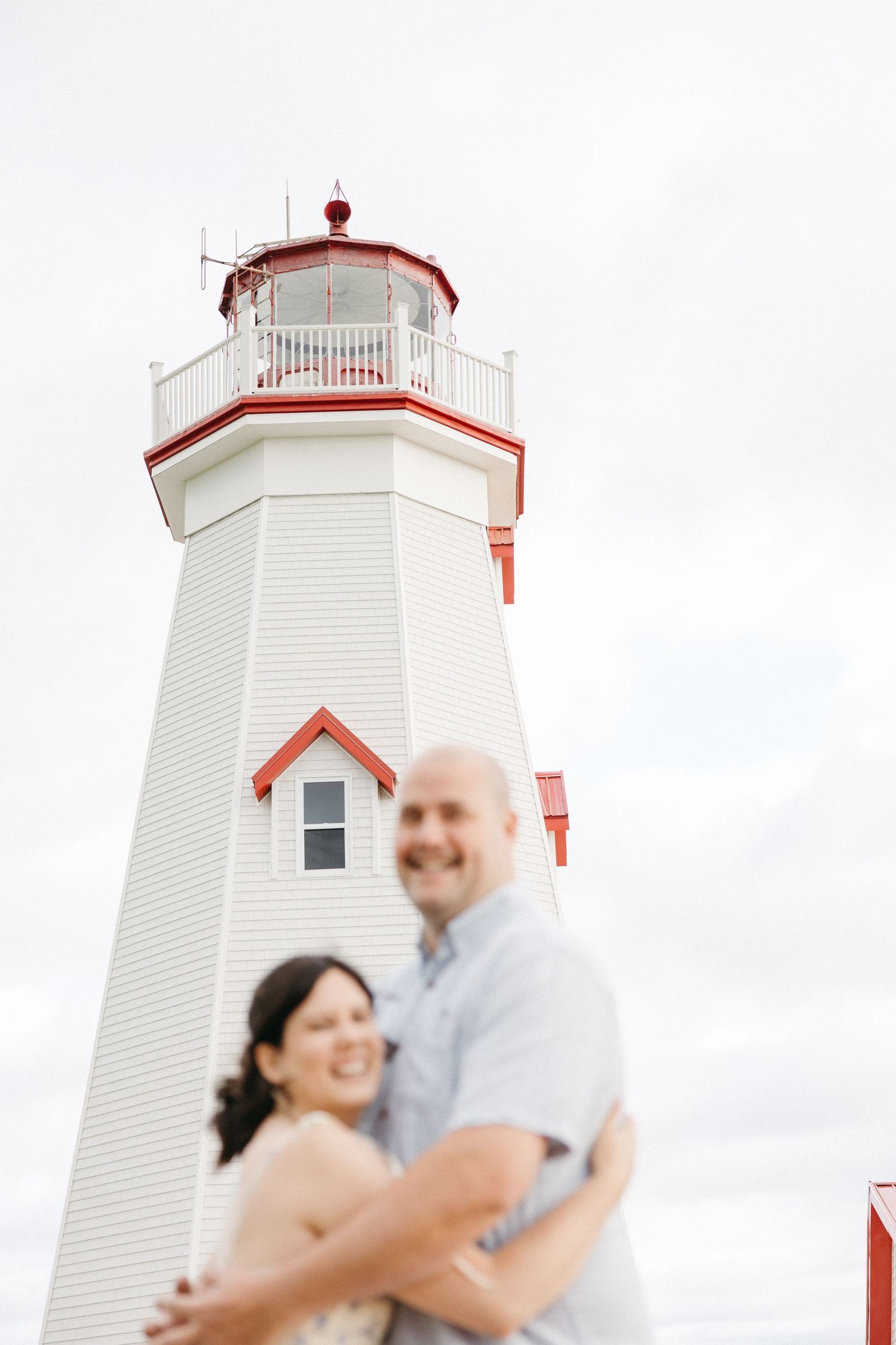 Smiling couple embracing in front of a white and red lighthouse under a cloudy sky.