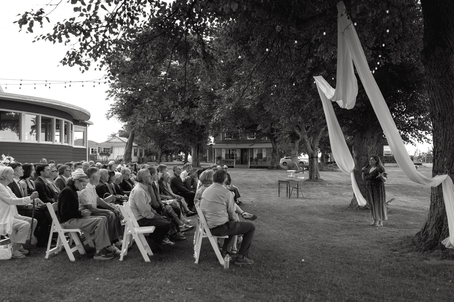 Black and white photo of wedding guests seated outdoors during a ceremony with elegant white fabric draped from trees.