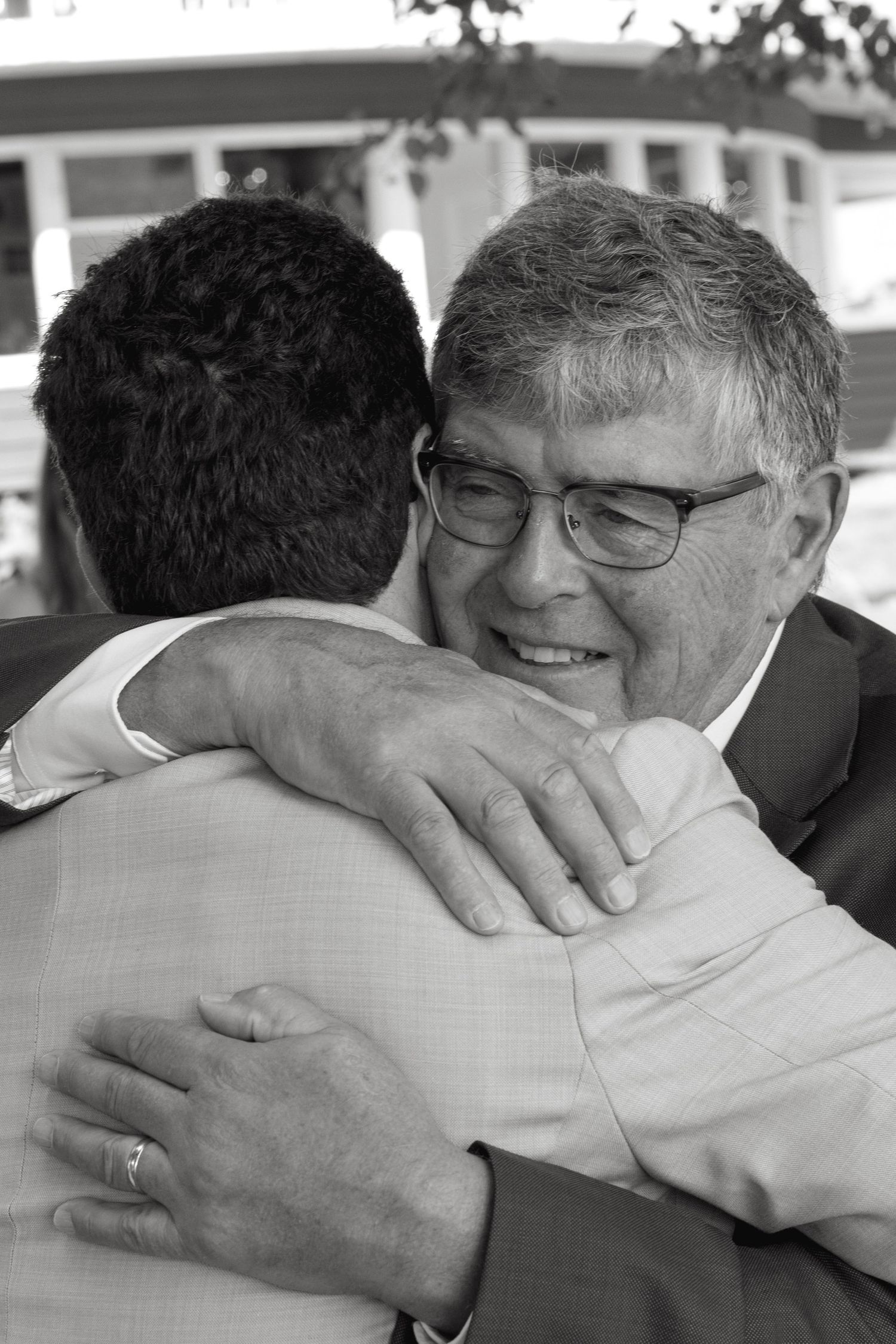 Older man in suit smiles warmly while embracing younger man in black and white wedding photo.