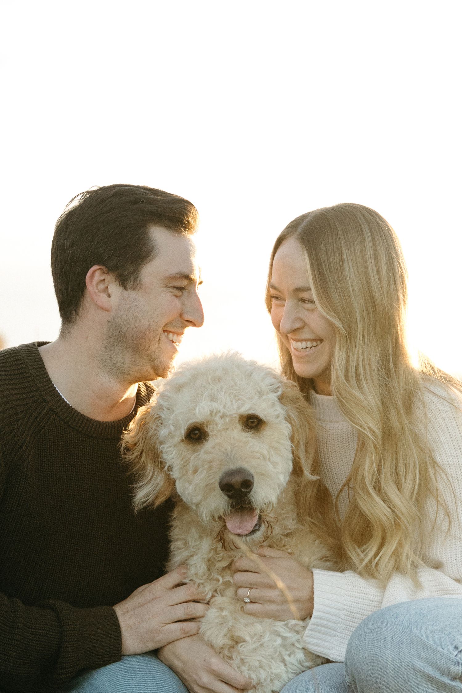 Smiling couple in cozy sweaters sit close together with a fluffy cream goldendoodle in warm golden sunset light.
