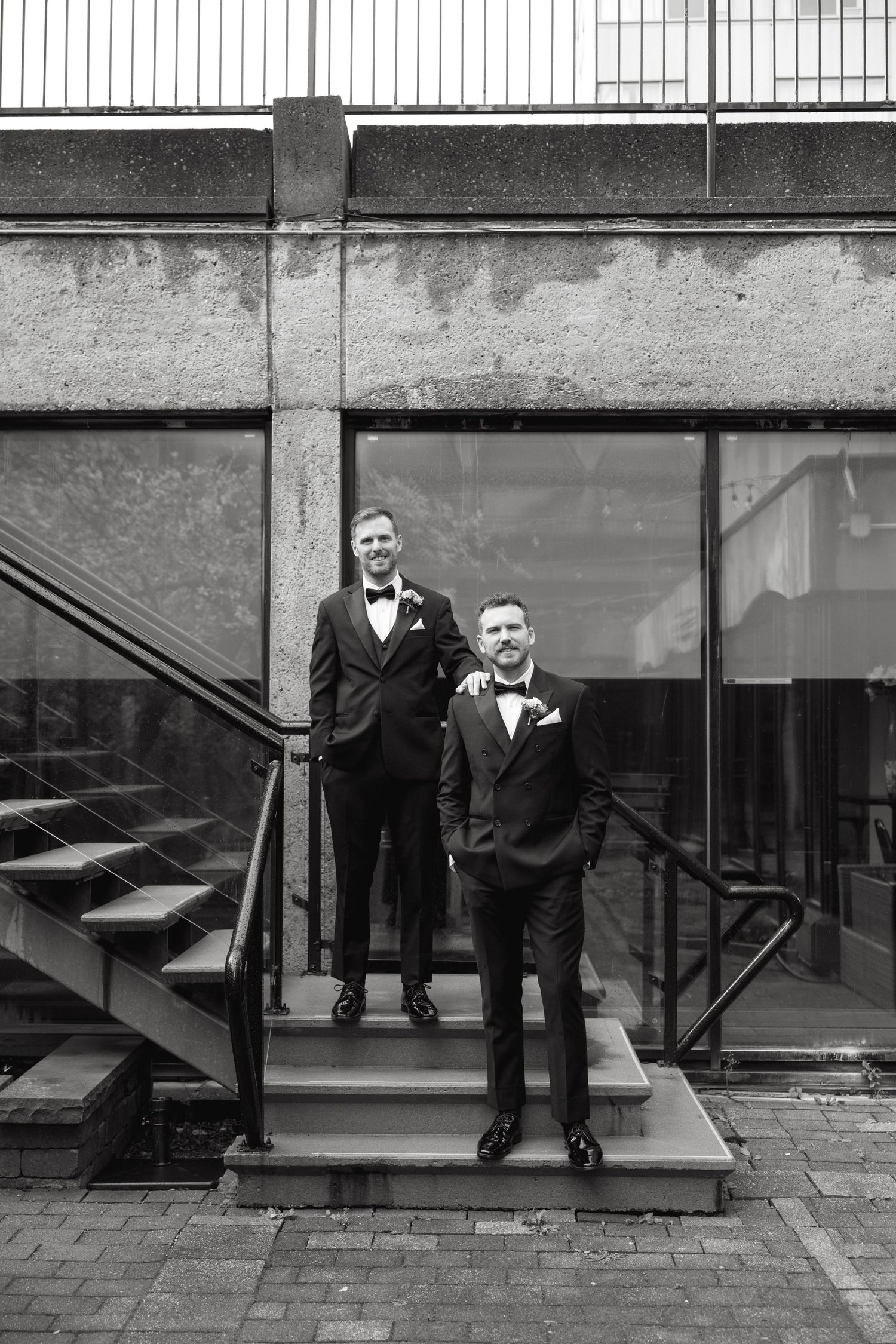 Two people in formal black tuxedos pose against an industrial backdrop.