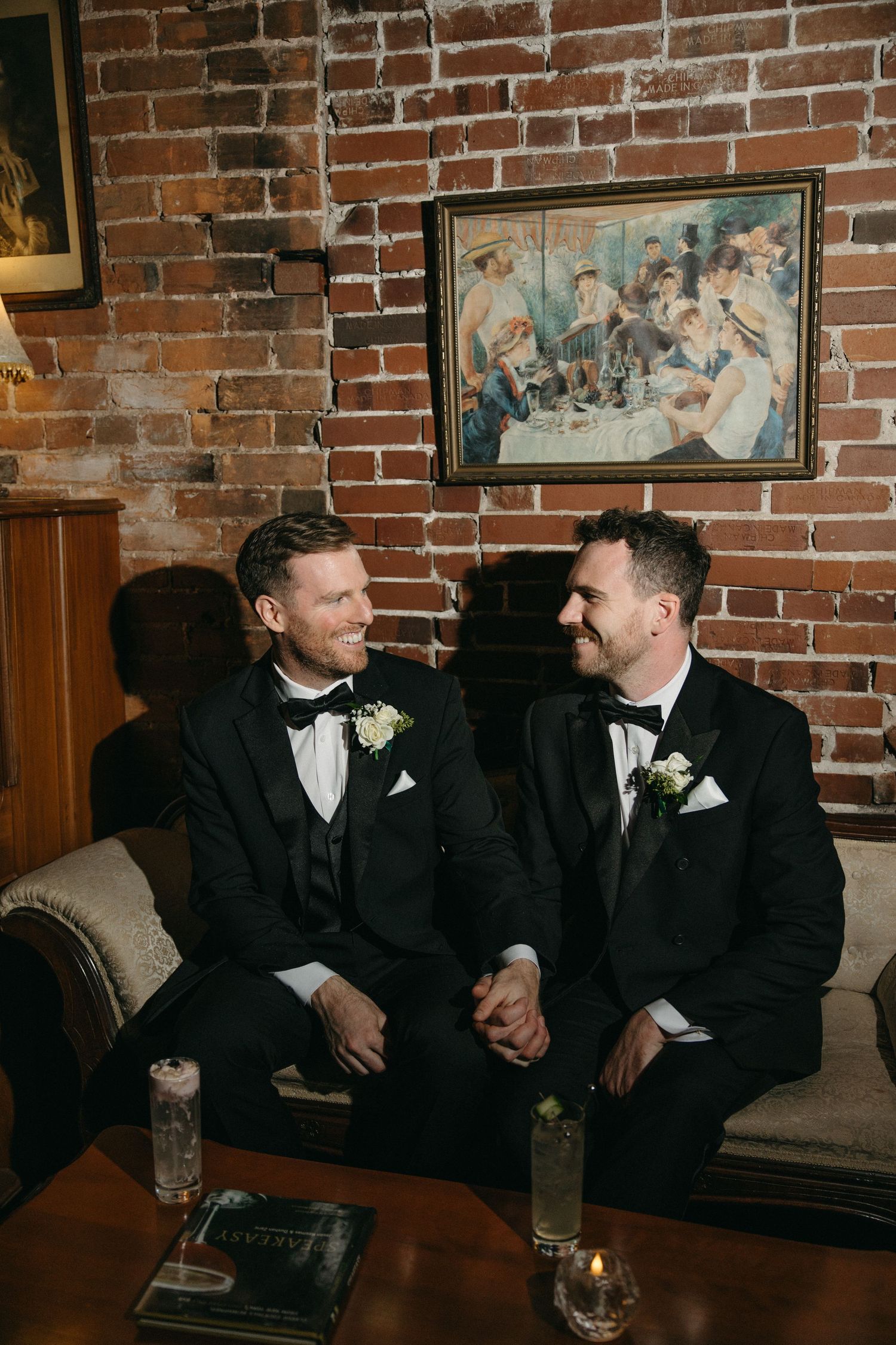 Groomsmen share laughs while seated on an elegant couch against rustic brick wall backdrop.
