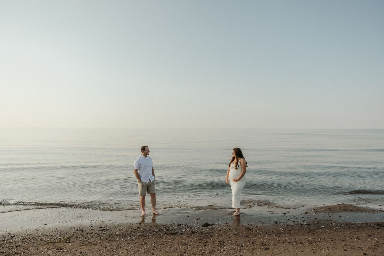 Two figures stand apart on a peaceful beach shoreline during golden hour.
