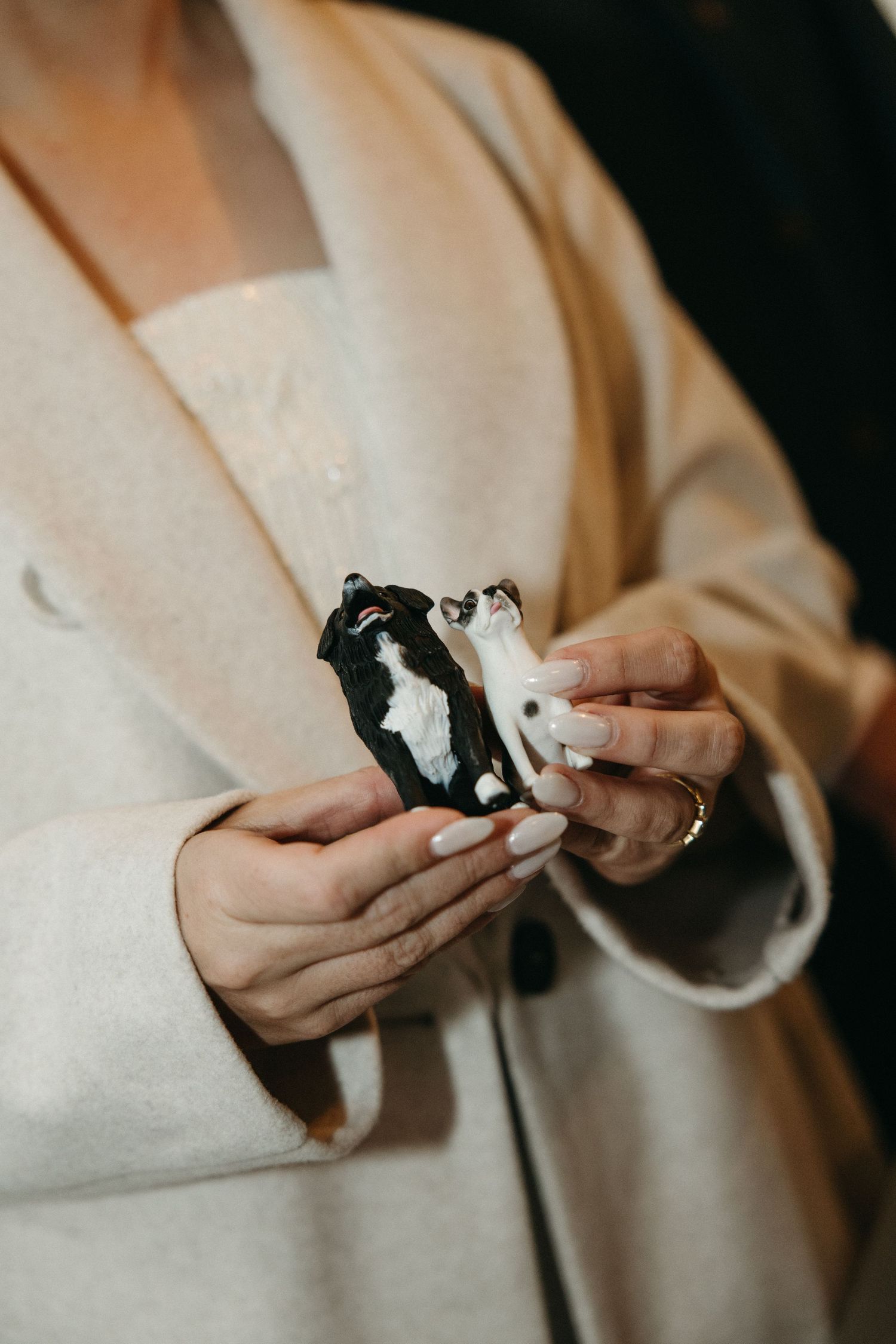 Woman in a cream wool coat with manicured nails holds two tiny black and white dog figurines, possibly wedding cake toppers.