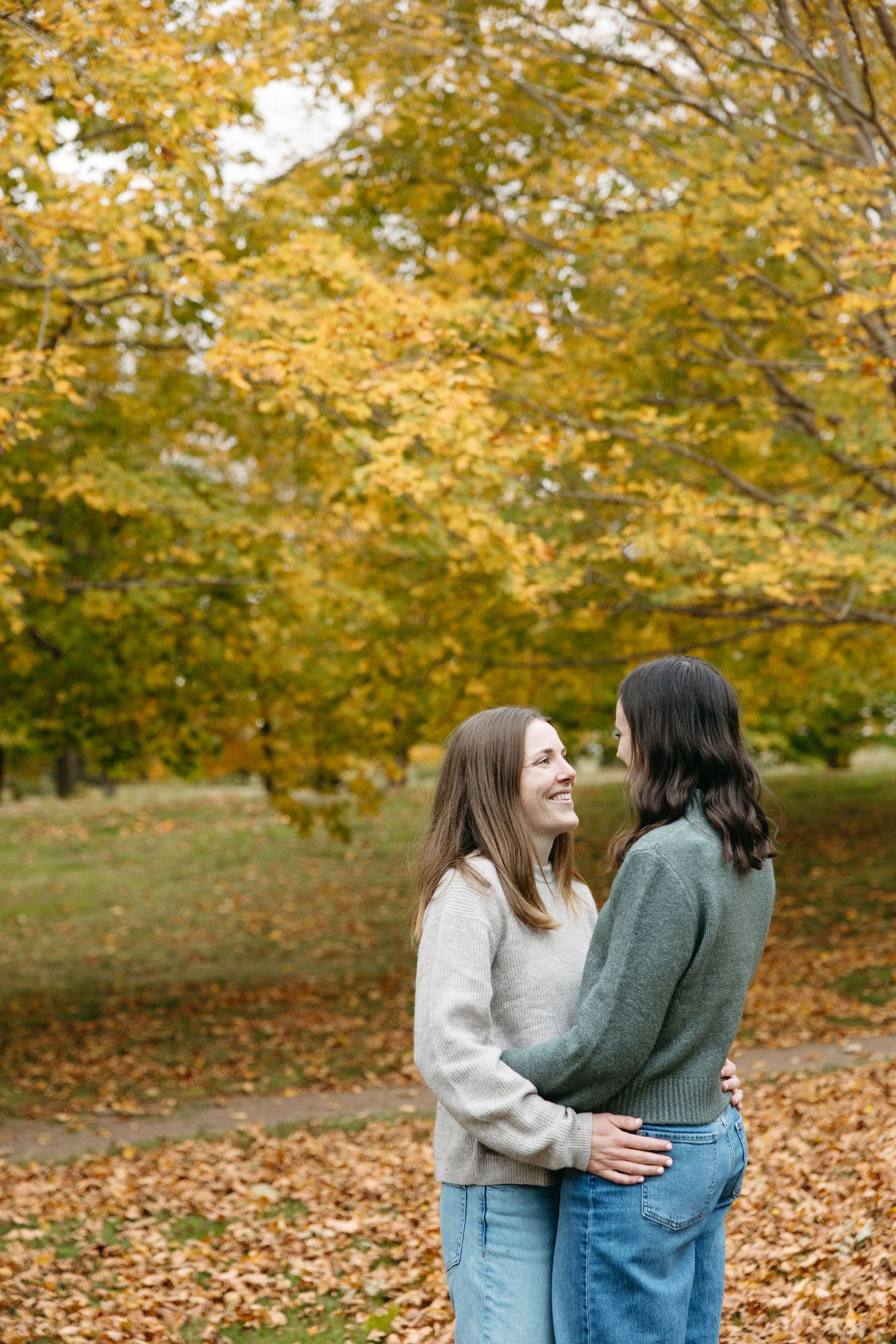 Two women embrace and smile in a park surrounded by golden autumn trees and fallen leaves, wearing cozy sweaters and jeans.