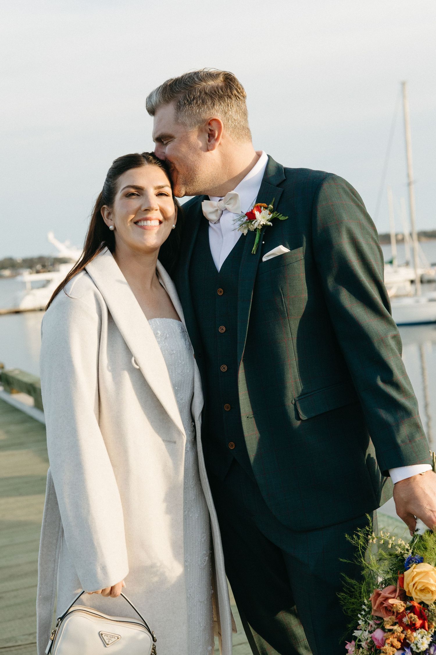 A groom in a dark green suit kisses the forehead of his smiling bride in a cream coat on a marina dock with sailboats behind them.