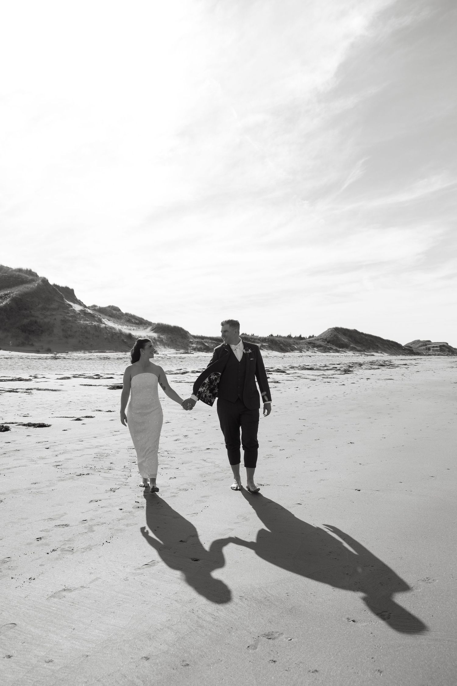 Black and white photo of a couple holding hands and walking barefoot along a sandy beach with dunes and long shadows.