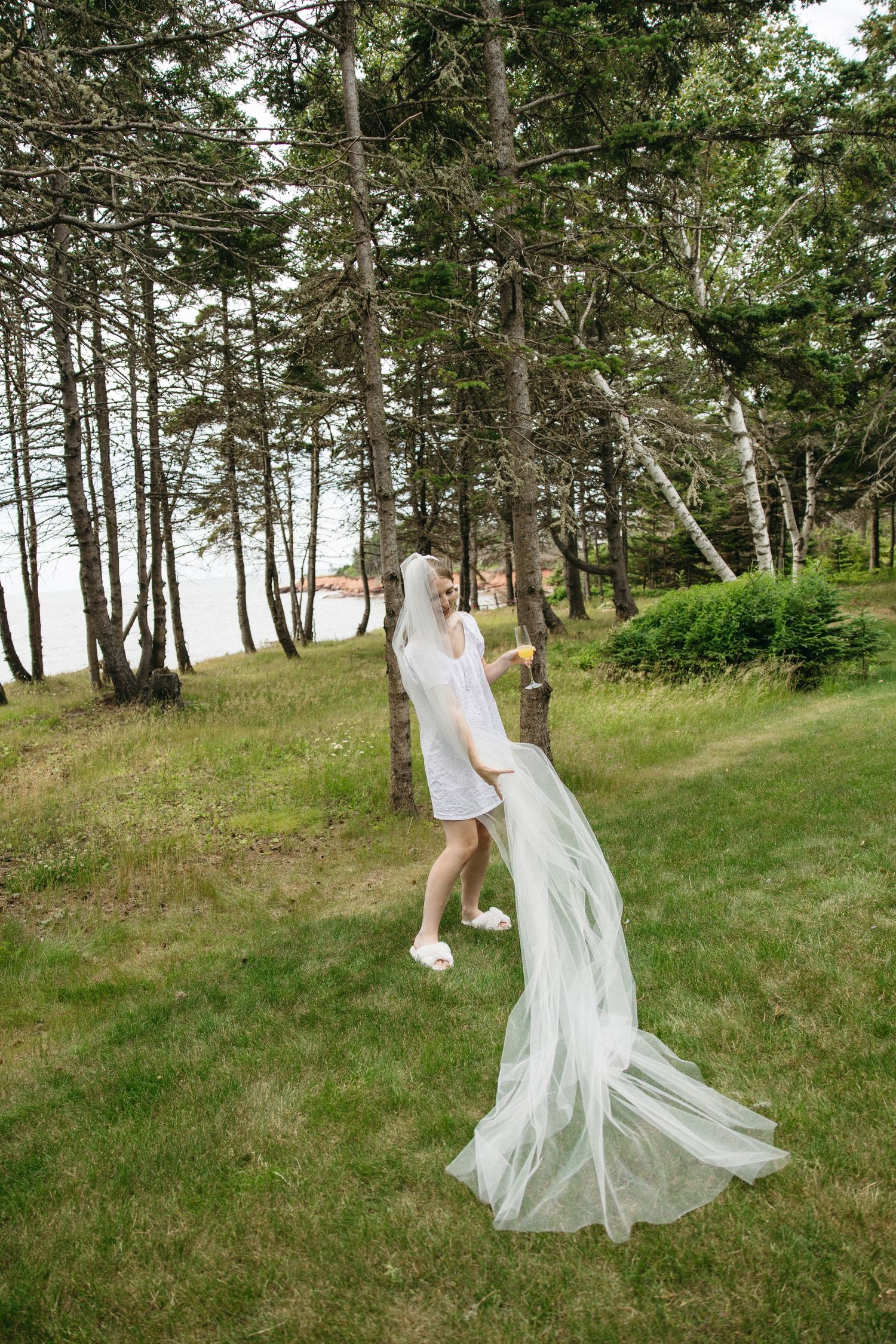 Bride in white dress and veil twirling outdoors near pine trees by the water on her wedding day.