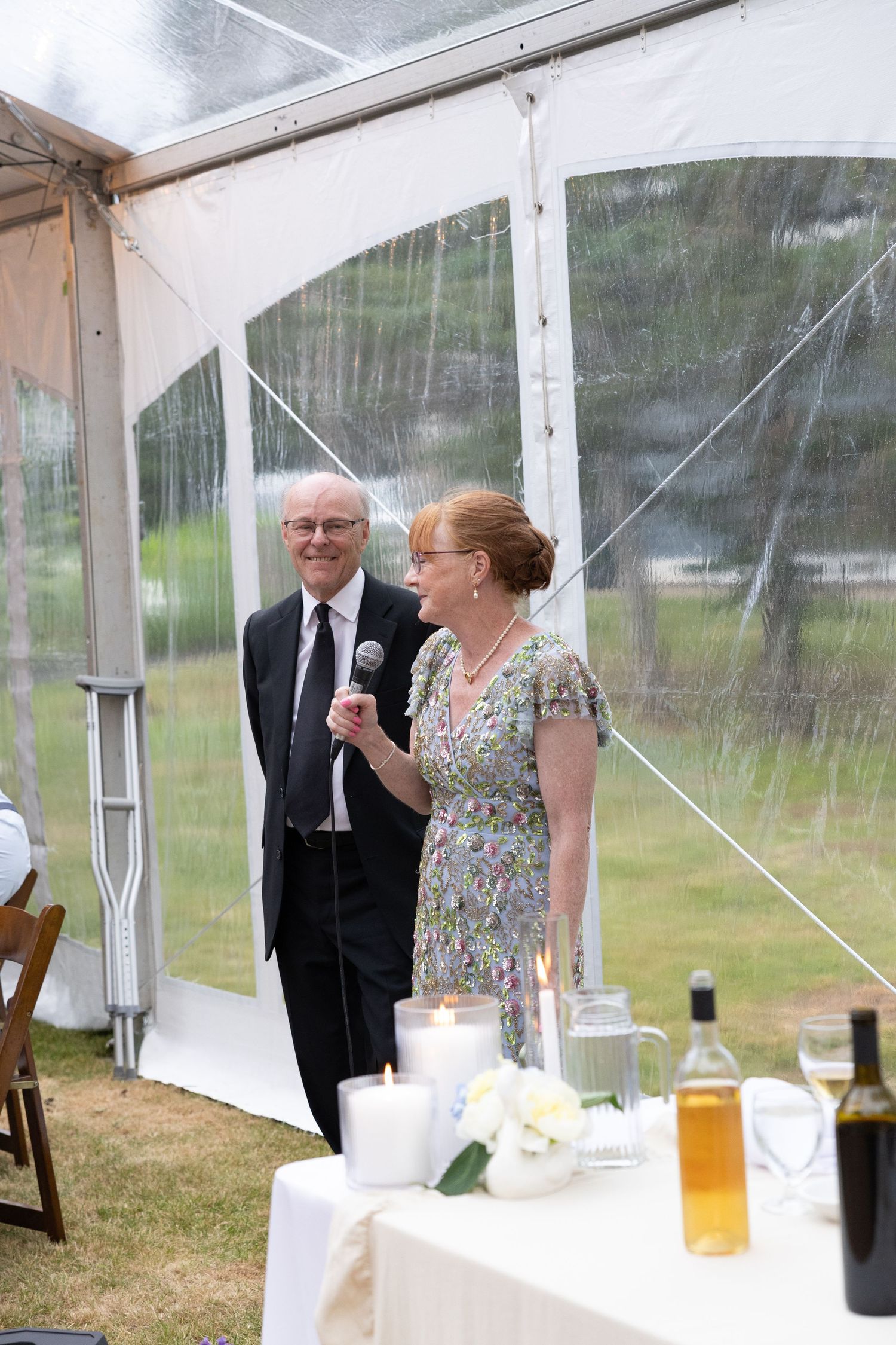 Older couple delivers wedding toast with microphone inside clear tent reception with candles and wine bottles.