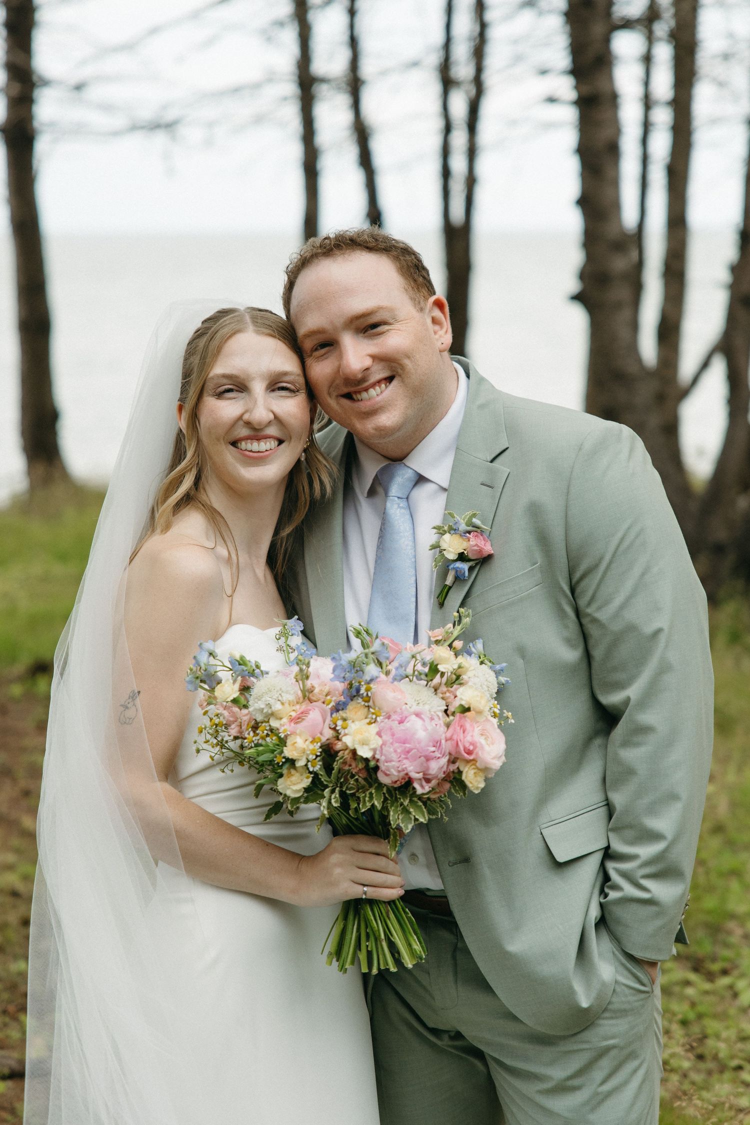 Smiling bride and groom pose together outdoors holding a pastel bouquet, groom in sage suit with blue tie.
