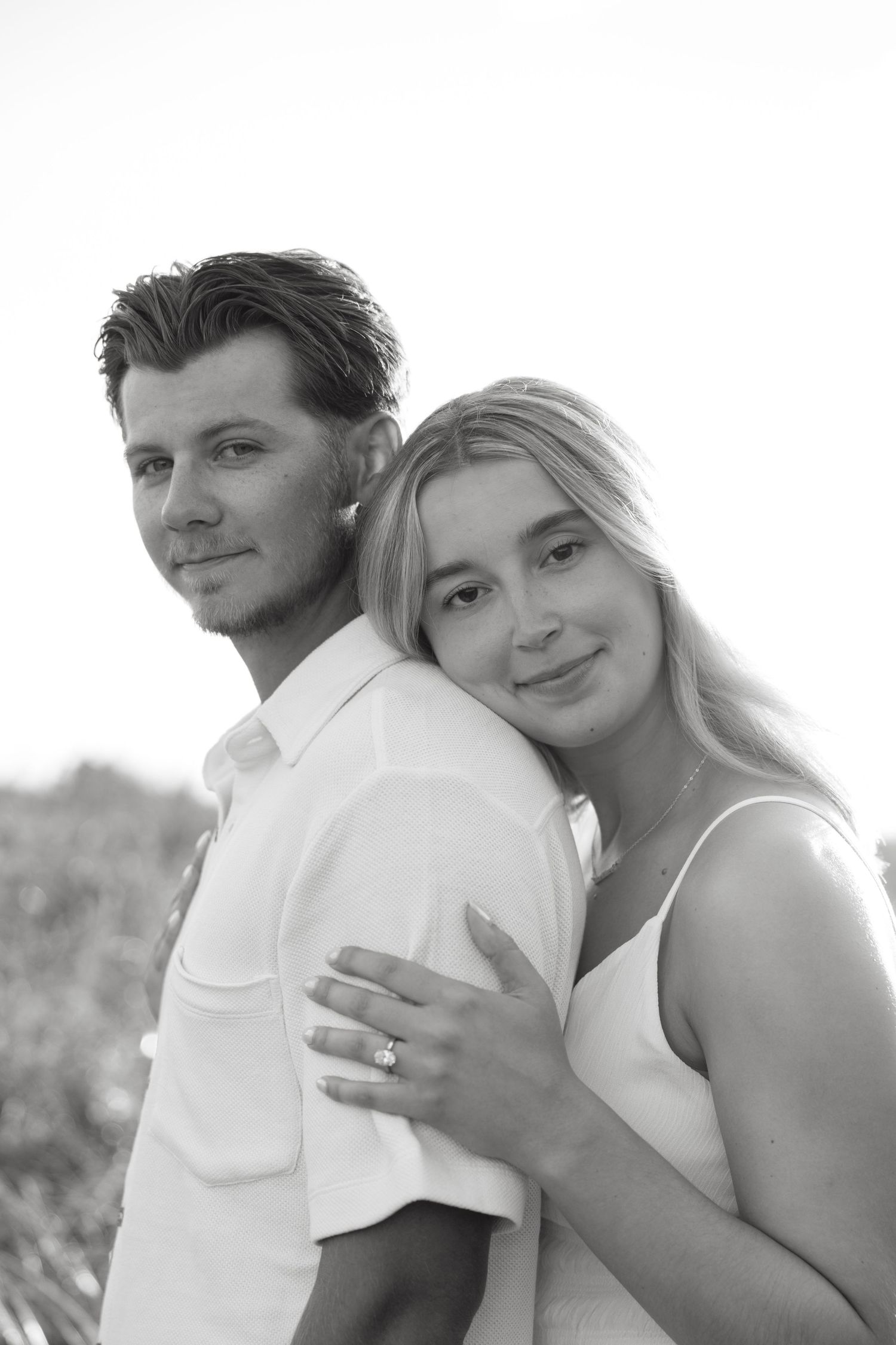 Black and white portrait of smiling couple, woman showing engagement ring outdoors.