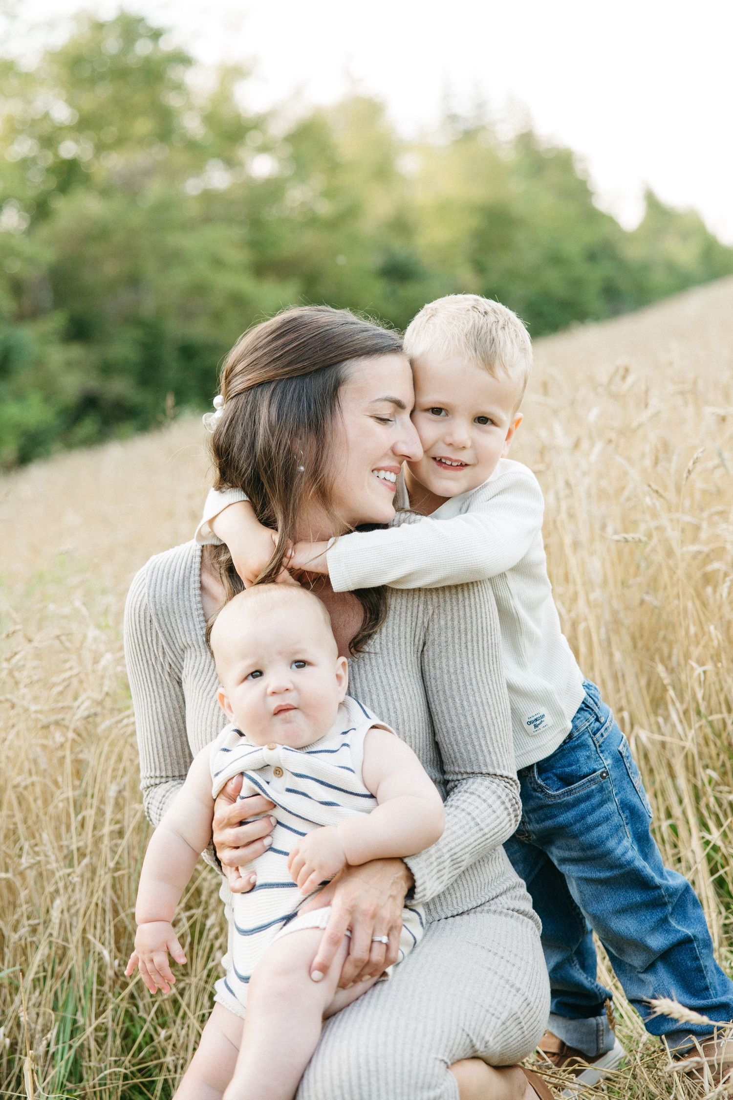 A mother kneels in a golden wheat field, holding her baby while her toddler son hugs her from behind.