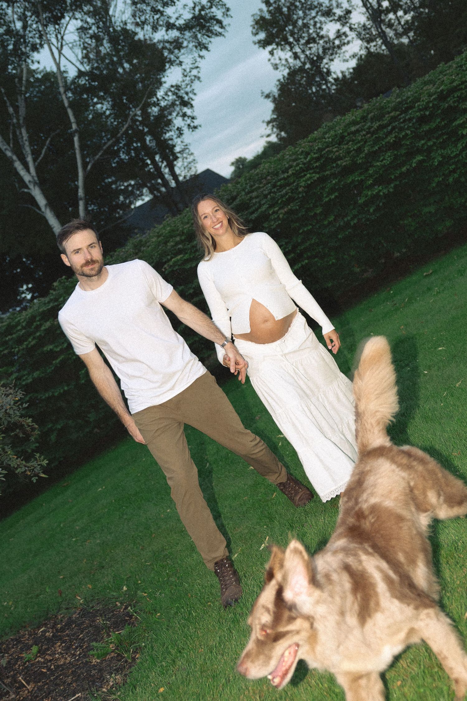 Smiling pregnant couple in white holding hands on green lawn while playful dog runs in foreground.