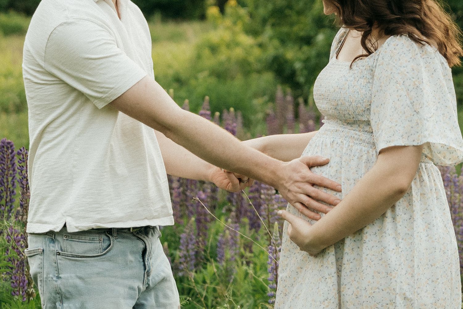 A couple in white outfits holds hands tenderly while standing in a blooming wildflower field on a sunny day.