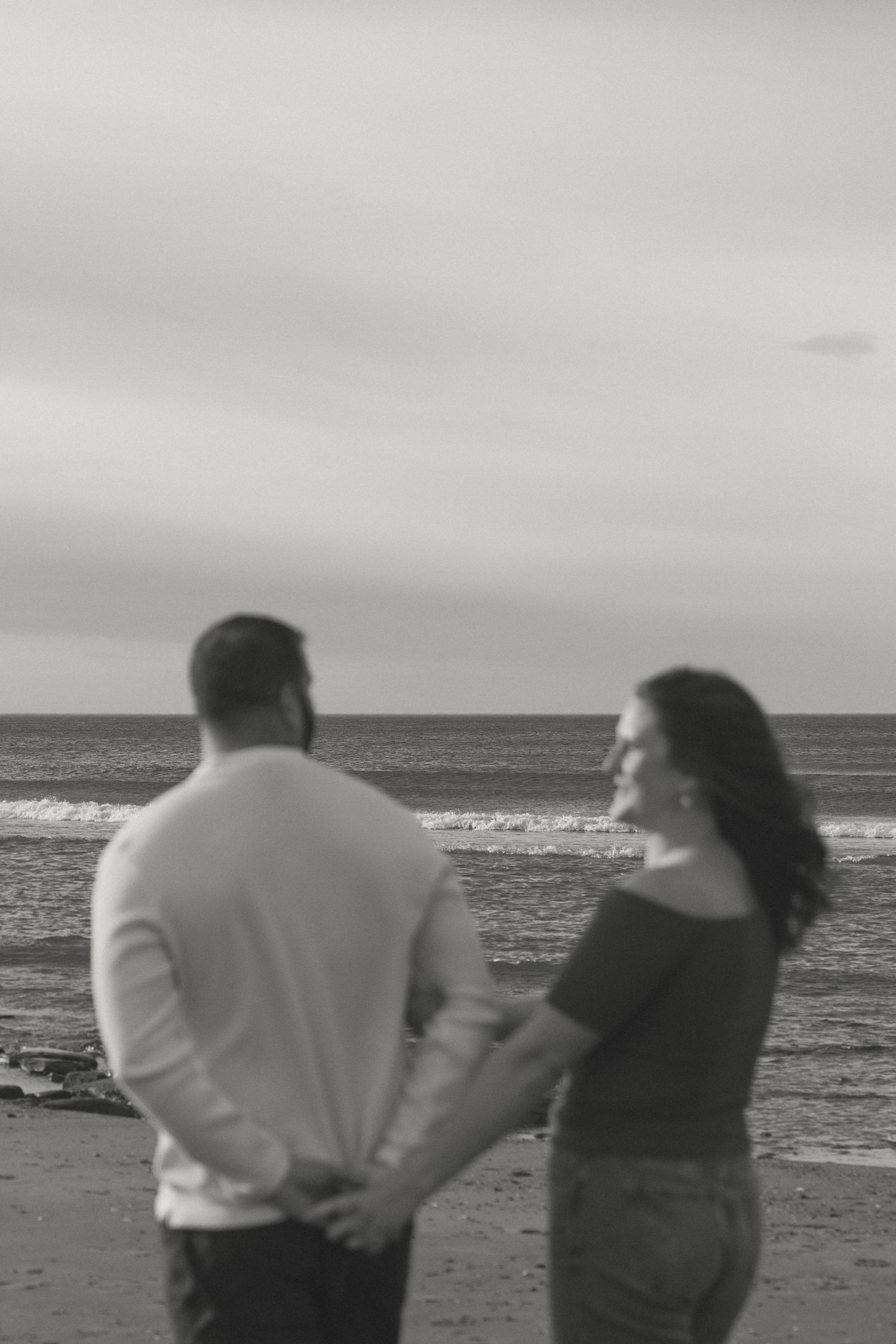 Black and white photo of a couple walking together on a beach with ocean waves in the background.