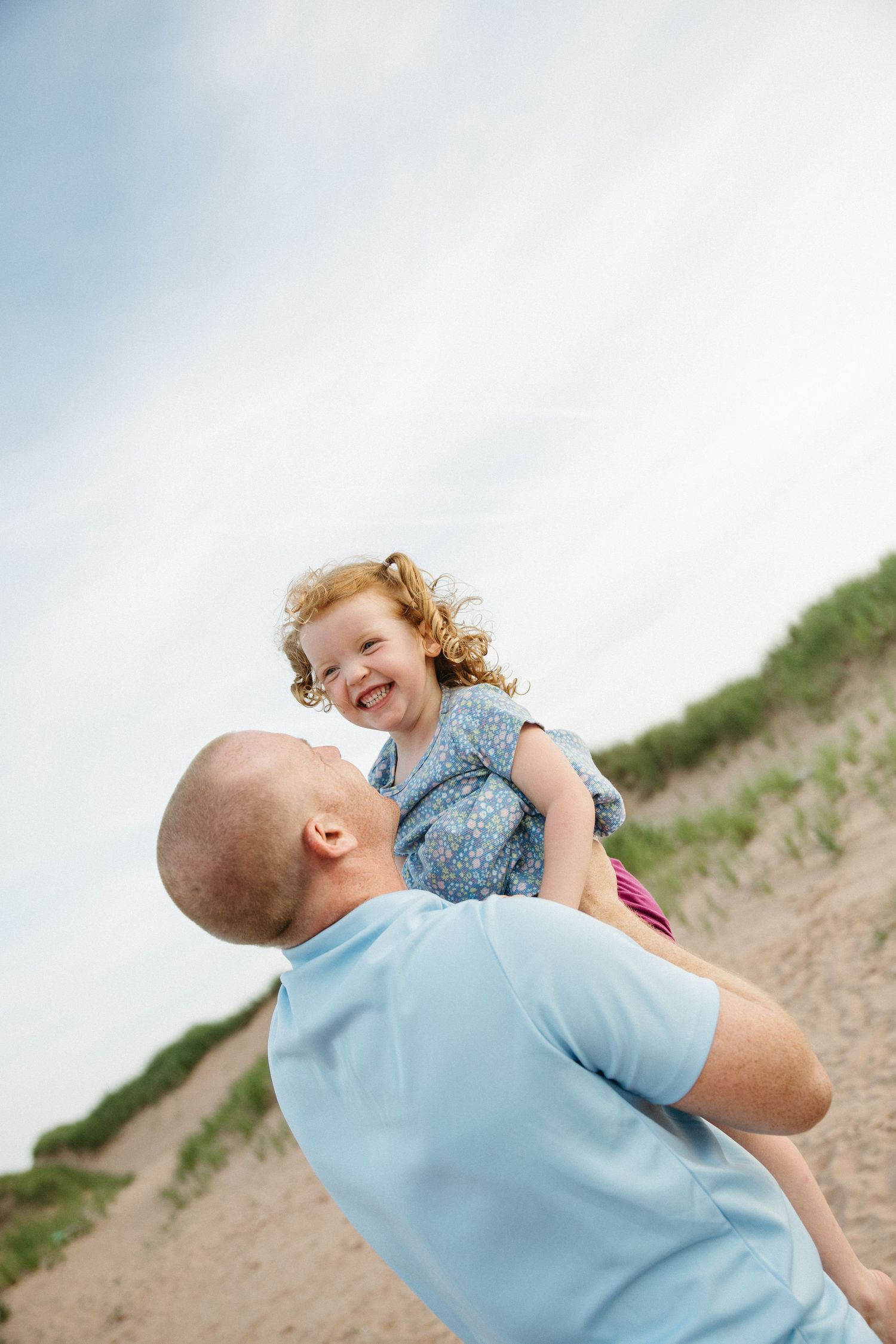 A father lifts his laughing curly-haired daughter on a sandy beach with grass dunes.