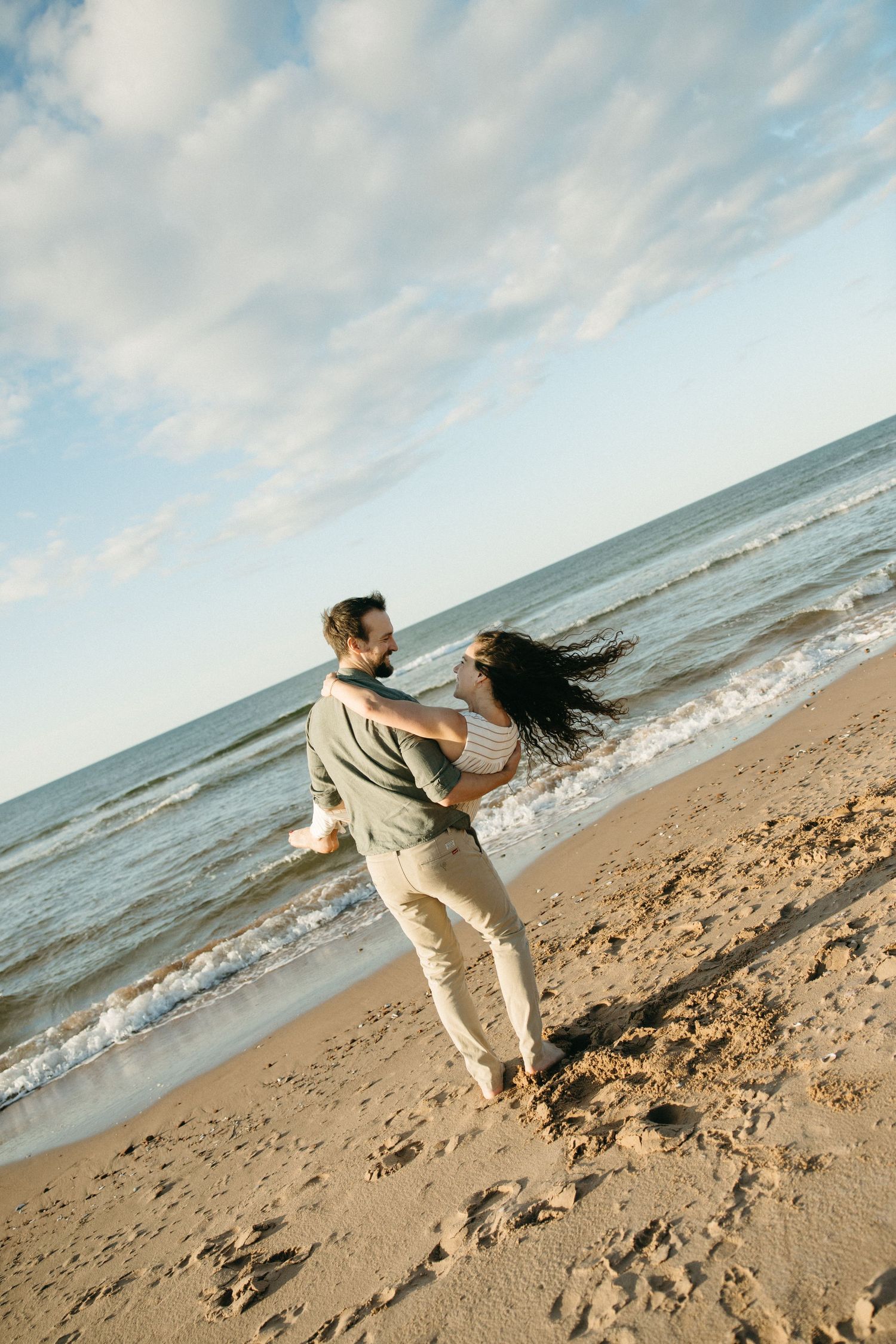 Couple embracing and dancing on a sandy beach with ocean waves and cloudy sky backdrop.