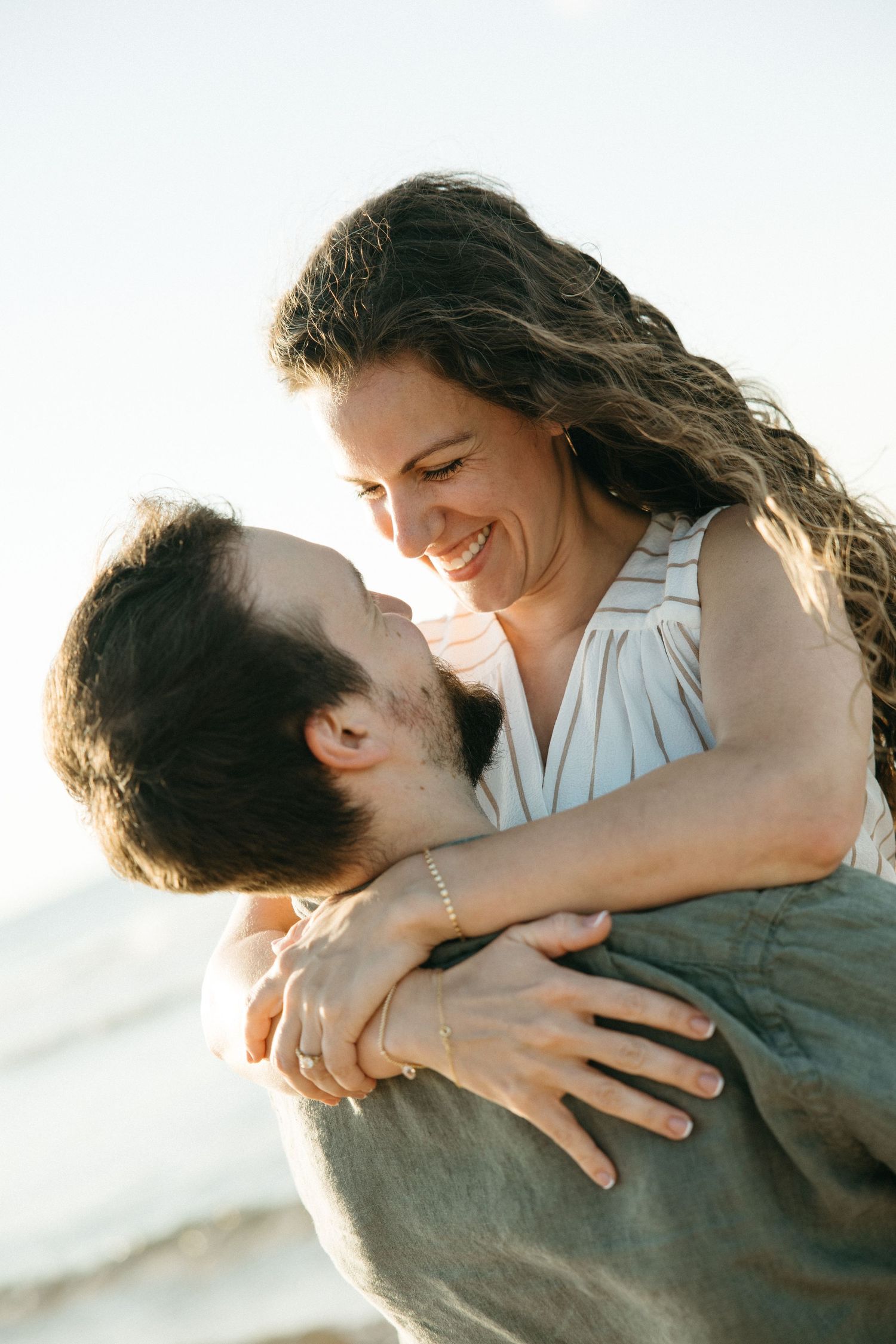 Happy couple embracing and smiling at each other on a sunny beach, woman wearing a ring.