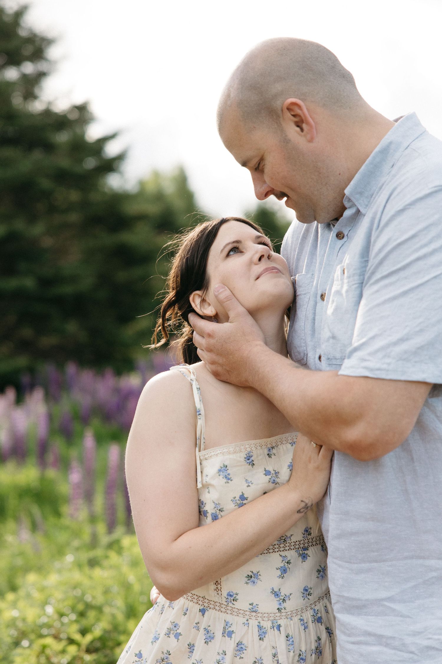 A couple shares a tender moment outdoors, man gently cupping womans face, with purple lupine flowers in background.