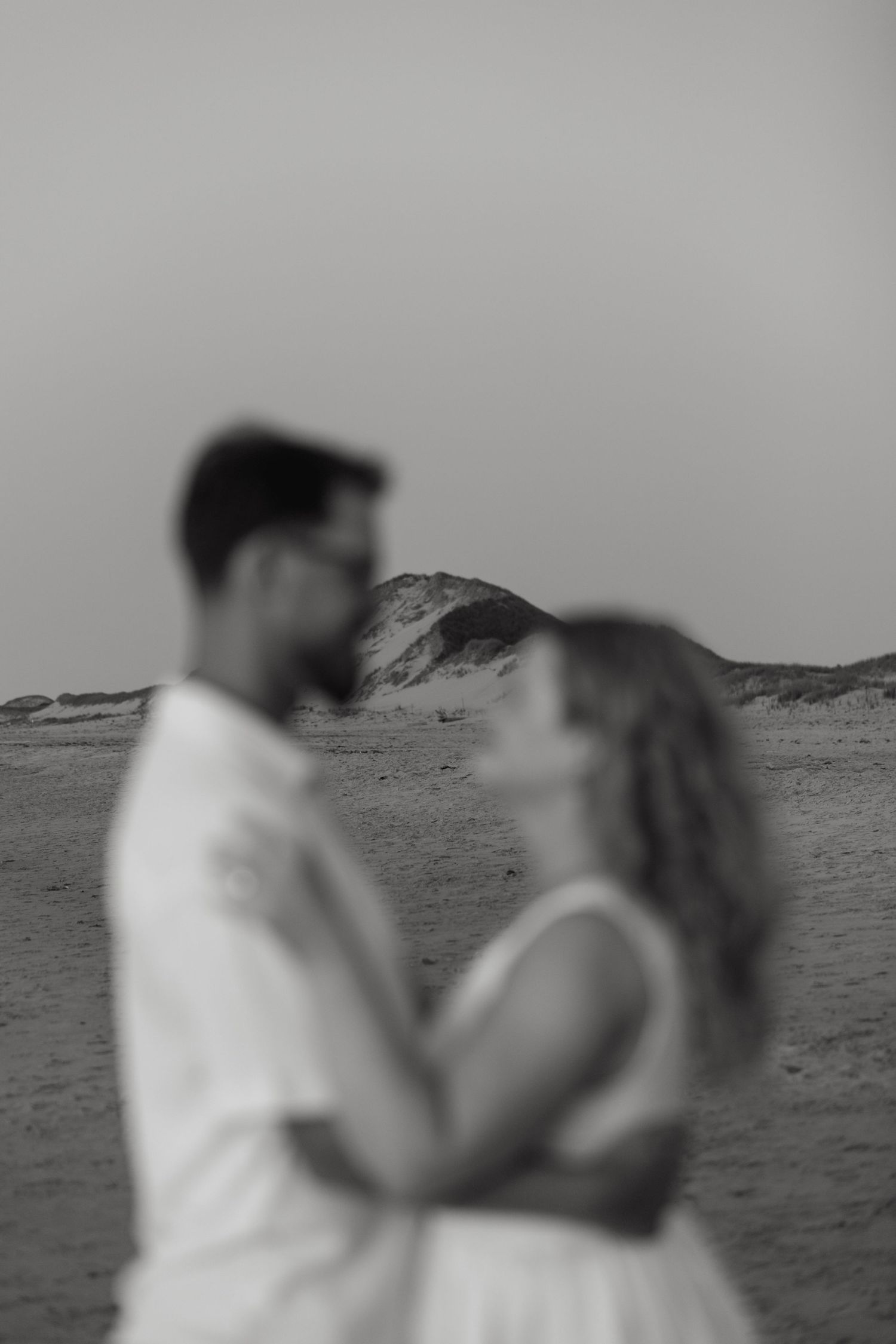 Black and white photo of a couple sharing a romantic moment on a sandy beach with dunes in the background.