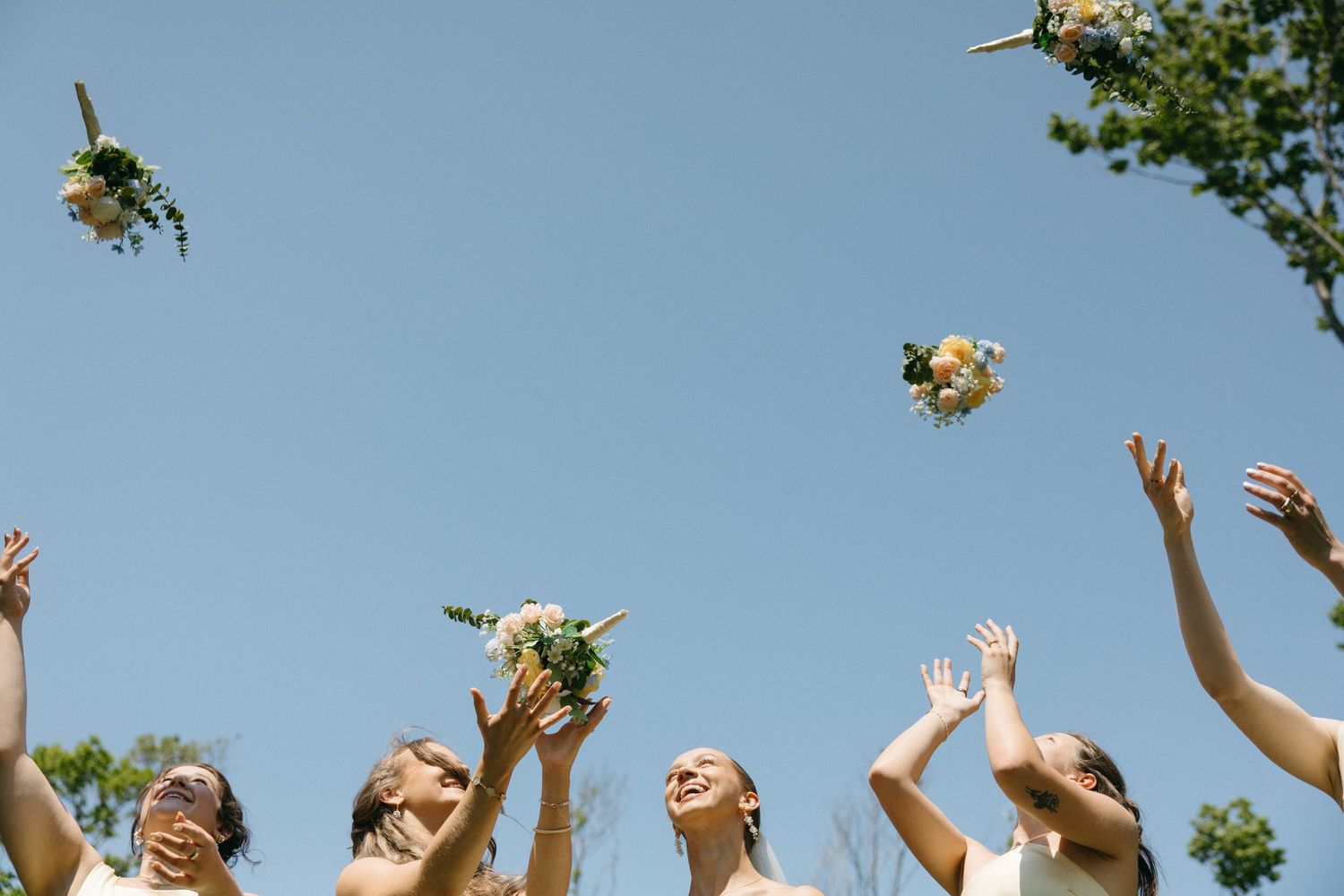 Bridesmaids in beige dresses joyfully toss floral bouquets into a clear blue sky outdoors.